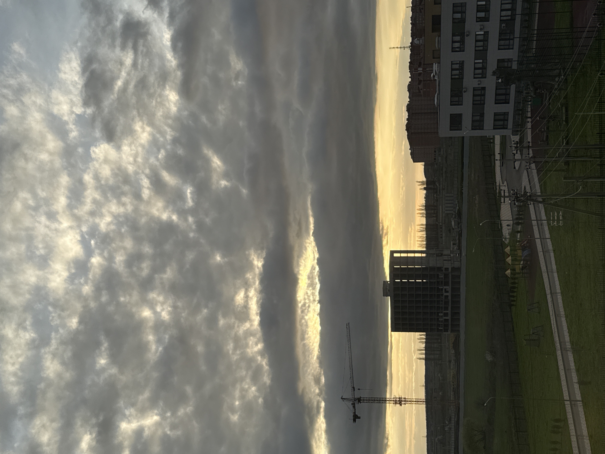 The image shows a view from an elevated position, possibly a building. The location appears to be an urban area during twilight. The sky is filled with textured clouds, with a strip of golden light visible, suggesting the sun is setting or rising. In the foreground, there is a construction site. A tall crane is visible, and there is a building under construction with a grid-like structure. To the right, there is a more established building with windows, appearing residential. A patch of green space is present, with what appear to be walking paths, streetlights, and potentially a playground. Power lines can be seen along the road. The image seems to capture a scene where development and nature coexist in the outskirts of a city.