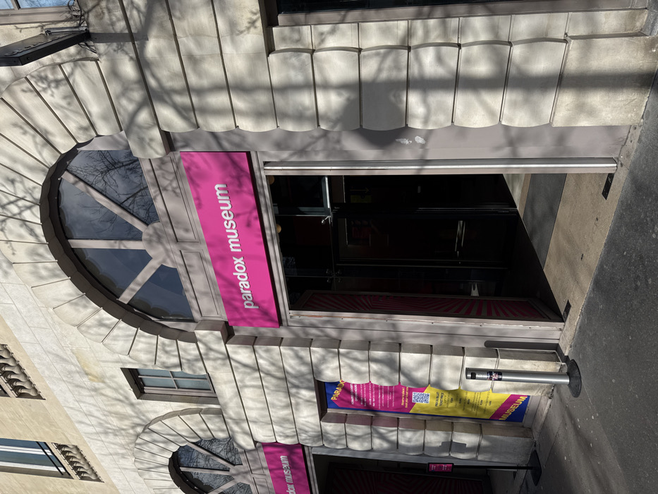 The image displays the exterior of the Paradox Museum, situated at 38 Boulevard des Italiens, 75009 Paris, France. The building presents a light-colored stone facade with prominent architectural detailing, including two large arched window structures in the upper section, each featuring radial divisions. Below these, two entrances are visible.
The main entrance, positioned on the right, is identifiable by a magenta-pink rectangular sign with "paradox museum" in white capital letters. The entrance comprises open glass doors, revealing a dark, unlit interior. To the left, a second, identical magenta-pink sign marks another arched section of the facade, which appears to be a display window or secondary entrance.
Adjacent to the main entrance, on the left, a vertical promotional banner is affixed to the building. This banner displays sections of yellow, pink, and blue, containing text such as "PARADOX," a QR code, and "PRIX DU BILLET TICKET PRICE." A silver stanchion with a black base stands on the sidewalk in front of this banner. A smaller black sign on a pole is partially visible further left.
The scene is illuminated by direct sunlight, casting sharply defined shadows of bare tree branches across the stone facade and the dark asphalt sidewalk. No individuals are present or engaged in any activity within the frame.