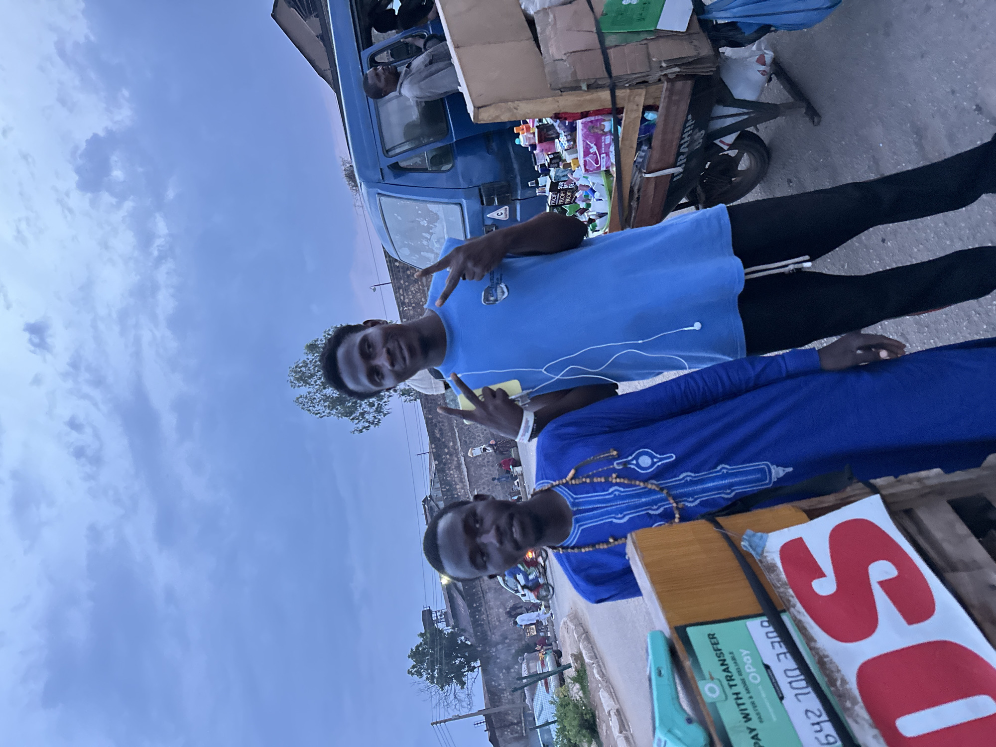 In Bukuru, Nigeria, a street scene unfolds under an overcast sky. Two young Black men are the central figures. The man on the left, facing the camera, wears a blue traditional tunic adorned with white embroidery and a beaded necklace. To his right, the second man, also Black, poses with his left hand raised in a peace sign, wearing a blue t-shirt and black pants, with white earbuds in.
Behind them, a blue van is parked, its driver visible through the window. Adjacent to the van is a cart laden with various goods, including bottles of what appear to be personal care products and packaged items. A wooden stand with a card reader and signage advertising "PAY WITH TRANSFER" is positioned in the foreground to the left. Partially visible red lettering on a white banner suggests a brand name.
The background reveals a street lined with buildings and a stone wall, hinting at the urban environment. A motorcycle and other vehicles are present on the road, and several other individuals are scattered throughout the scene, some appearing to be vendors or customers. The overall atmosphere suggests a busy marketplace or roadside commerce.