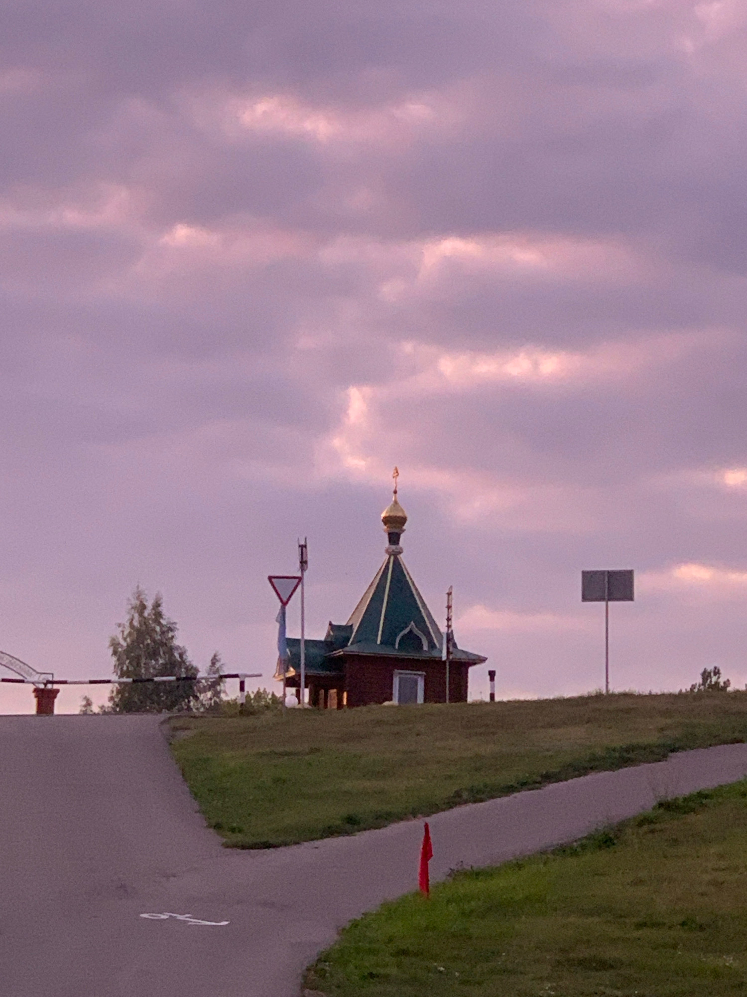The image depicts a small, red building with a green roof and a golden dome, resembling a chapel or a small church. It is situated on a grassy hill. In the foreground, a paved road curves to the left, with a white symbol resembling a cyclist painted on it. To the left of the road, a striped barrier is visible. Behind the chapel, there is a traffic sign and another sign post. The sky above is filled with pink and purple clouds. The scene appears to be an outdoor location, likely in a rural or semi-rural setting, given the presence of the road, grass, and the chapel. There are no people visible in the image. The overall atmosphere suggests dusk or dawn due to the lighting and cloud colors.