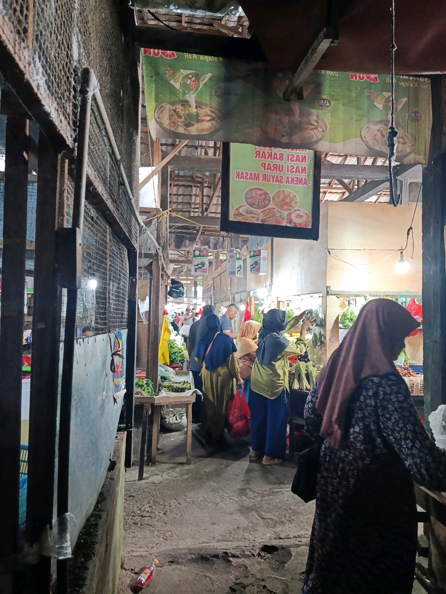 This image depicts a bustling marketplace in Petung, Indonesia, characterized by its rustic and somewhat makeshift architecture. The scene is dominated by stalls displaying various goods, with a focus on fresh produce. In the foreground, a woman wearing a patterned abaya and a pink hijab is partially visible from the back, seemingly engaged in shopping. Further into the market, several other individuals, predominantly women, are seen browsing and interacting with vendors. They are mostly dressed in traditional Islamic attire, including hijabs and abayas of various colors. Some are carrying shopping bags, including a red plastic bag. The stalls are constructed from wood and metal, with some covered by tarps or banners. One banner, prominently displayed, features images of food and Indonesian text. The ground is uneven and appears to be made of concrete. The overall impression is one of a vibrant local market, likely operating in the morning or afternoon, given the lighting. A bottle of what appears to be soy sauce or chili sauce is lying on the ground near a low concrete wall on the left side of the image.
