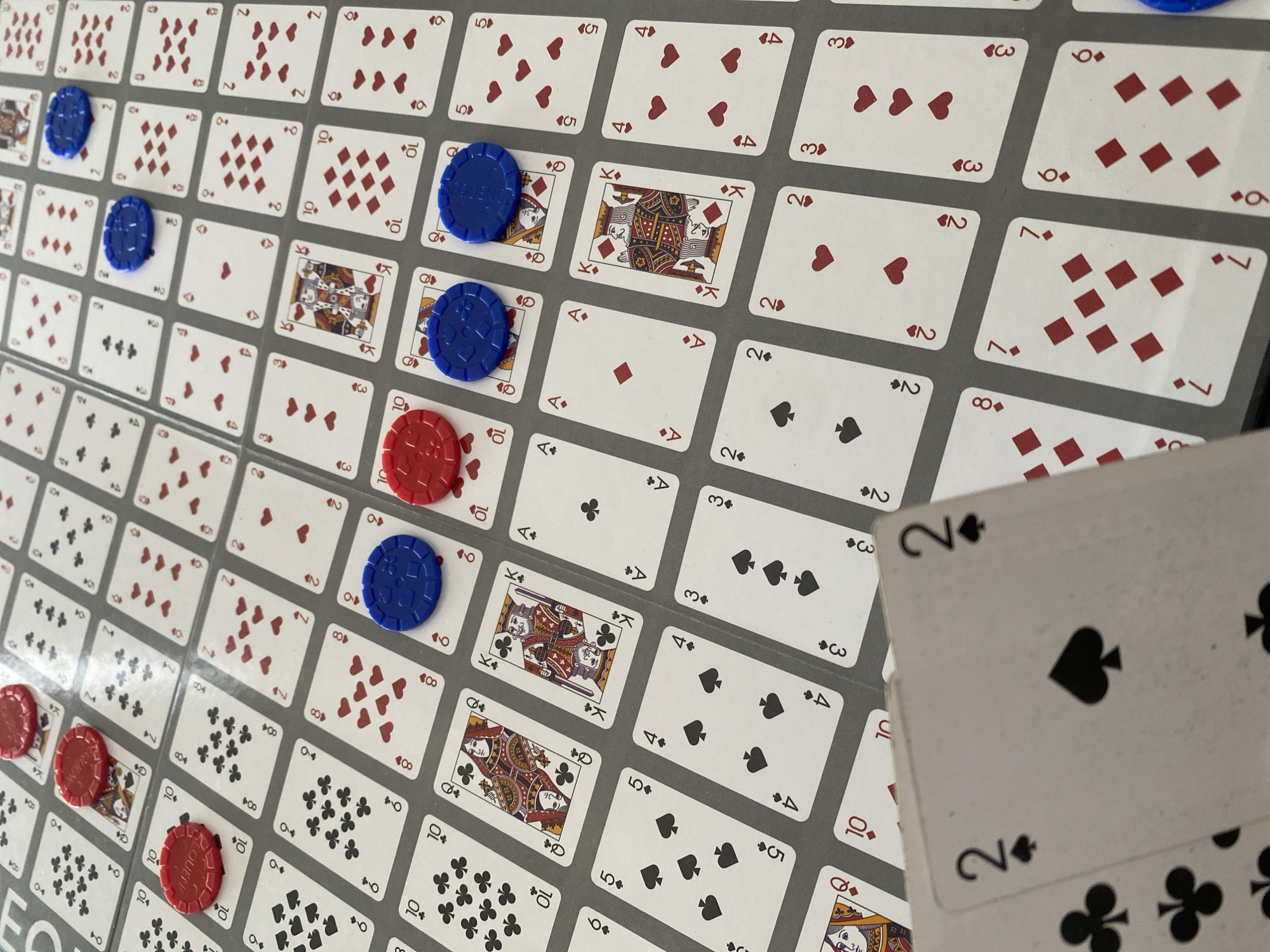 The image depicts a Sequence game board, filled with playing card images in a grid pattern. Several blue and red plastic chips are placed on some of the cards. Two playing cards, a 2 of Spades and a 2 of Clubs, are held in the foreground. The background is out of focus.