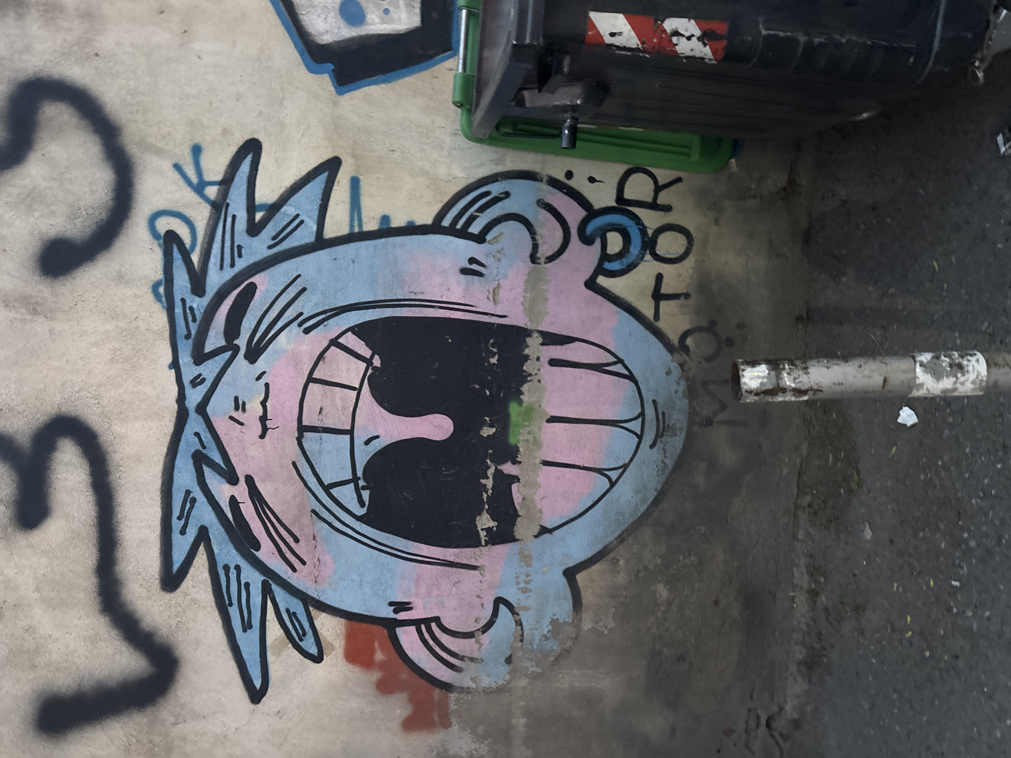 The media file shows a close-up view of a concrete wall with graffiti. The primary subject is a cartoonish drawing of a face, rendered in light blue and pink with black outlines. The face has a wide-open mouth, revealing sharp teeth and a black interior. Jagged blue lines emanate from the top of the head, resembling hair or spikes. To the left of the face, a large, dark, curved graffiti tag is visible.
Above the cartoon face, a portion of a dark green dumpster with a red and white striped sticker is visible. To the right of the face, the text "TOR" is spray-painted in black. The graffiti and the dumpster are situated on a concrete surface that appears to be outdoors, possibly a street or alley, as indicated by the adjacent asphalt ground. A metal pole, partially painted white, runs horizontally across the lower right portion of the image. The lighting suggests it is daytime, but no specific time or weather conditions can be determined. The overall impression is one of an urban environment with visible street art.