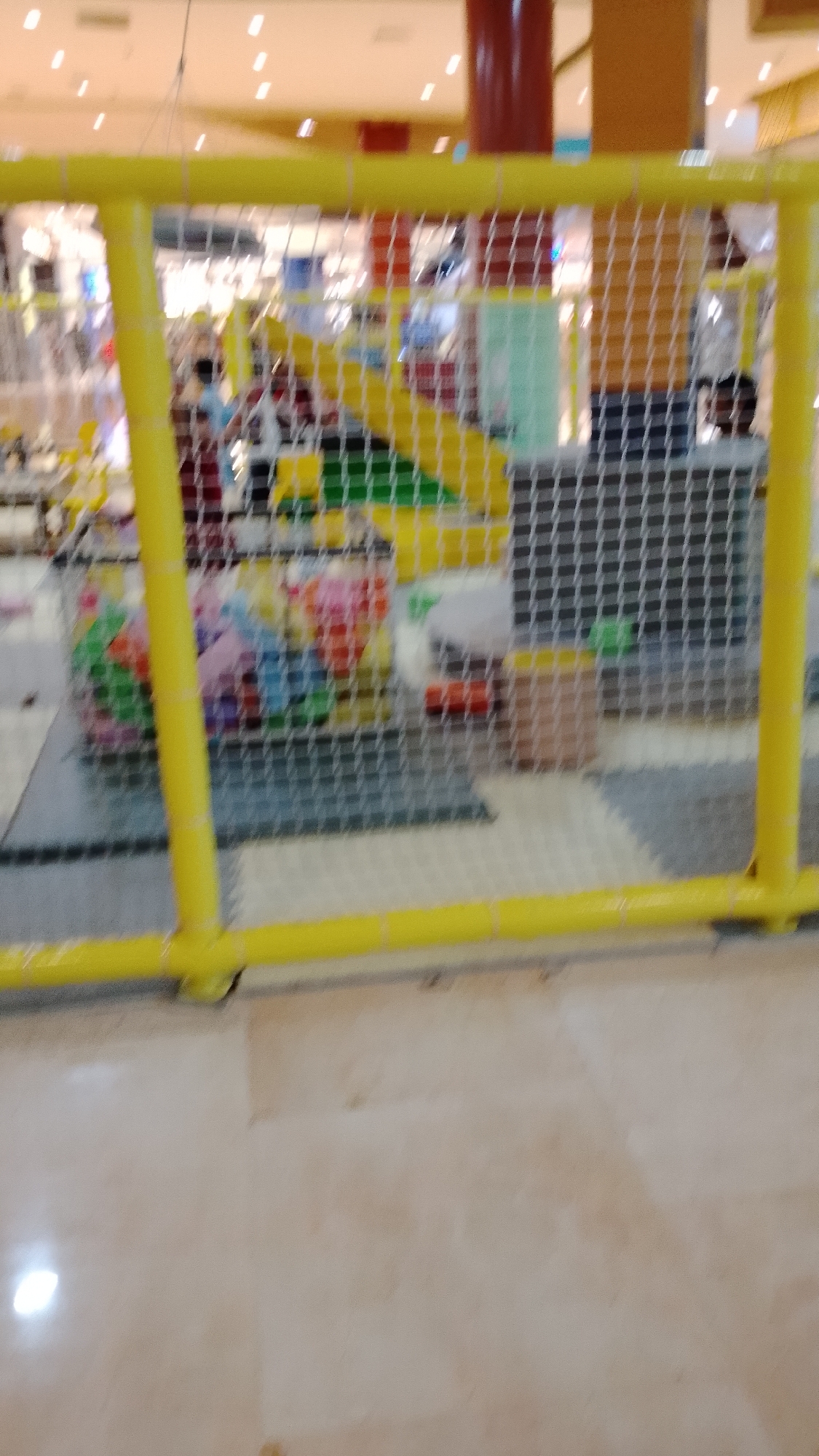 This image depicts a brightly colored indoor playground, likely located in South Tangerang, Indonesia, as suggested by the context. The foreground is dominated by a yellow tubular safety fence, part of the playground's structure, with a mesh netting extending behind it. Through the netting, a ball pit filled with multi-colored plastic balls is visible. Further in the background, hints of play structures like slides and climbing areas can be discerned. The overall lighting suggests an indoor environment with overhead lighting, and some of the background elements are blurred due to the focus on the foreground and the image's resolution. A person's head is partially visible in the far right background, looking towards the left. The floor appears to be made of soft, interlocking mats.