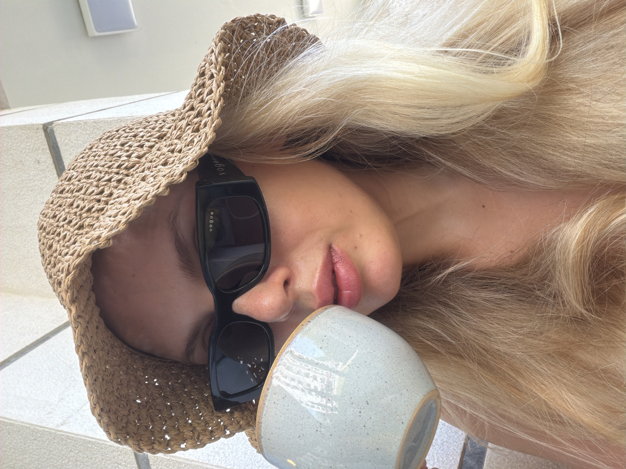 In Budva, Montenegro, a blonde woman wearing a woven straw hat and black sunglasses is captured in a close-up shot as she sips from a light blue ceramic mug. The mug, with a yellow rim, reflects the surrounding environment. Her lips are slightly parted as she drinks. The background consists of a light-colored textured wall, possibly part of an outdoor seating area. Her long, blonde hair is visible, framing her face and falling over her shoulders. The image has a casual, relaxed feel, suggesting a moment of leisure.