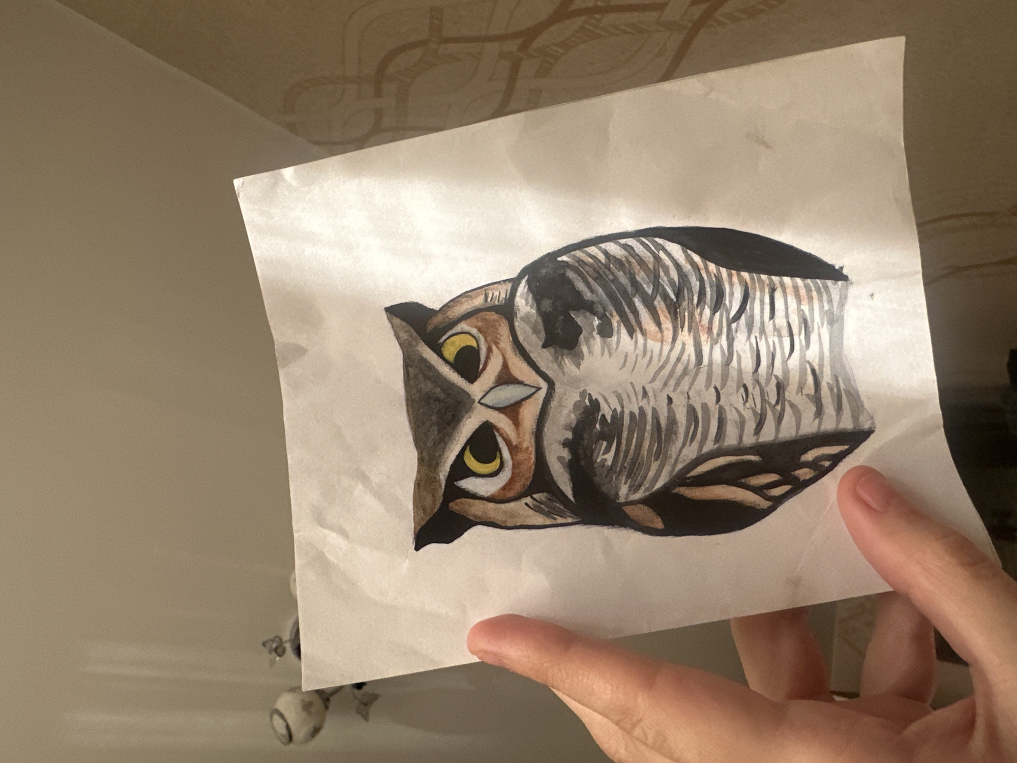 An indoor photograph captures a right hand holding a white piece of paper featuring a drawing of an owl. The owl, drawn horizontally on the vertically held paper, is depicted in shades of brown, grey, black, and white, with prominent yellow eyes. Its feathers are rendered with distinct strokes, suggesting a watercolor or marker medium. The paper itself shows subtle creases and wrinkles. The hand grips the bottom-right corner of the paper, with visible fingers and fingernails.
The background consists of a plain, light beige-brown wall in the mid-ground. Above the wall, a ceiling with a subtle, intricate patterned relief or wallpaper in a similar muted tone is visible, featuring intertwining, circular designs. Below the paper, partially obscured, is a small, light-colored object with rounded components, possibly a decorative item or a toy. The overall lighting is soft, suggesting an indoor setting. There is no visible text in the image. The scene is consistent with an interior setting in Uva, Russia.