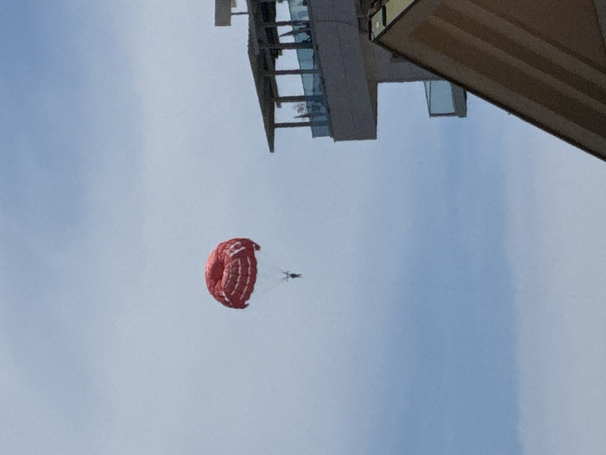 A parasailer with a red parachute is visible in the sky over Budva, Montenegro. The parasailer is suspended below the parachute by ropes. In the foreground, a portion of a modern building with balconies is visible, along with another building structure that appears to be a rooftop or upper floor with a slanted roofline. The sky is overcast with light blue and white clouds.