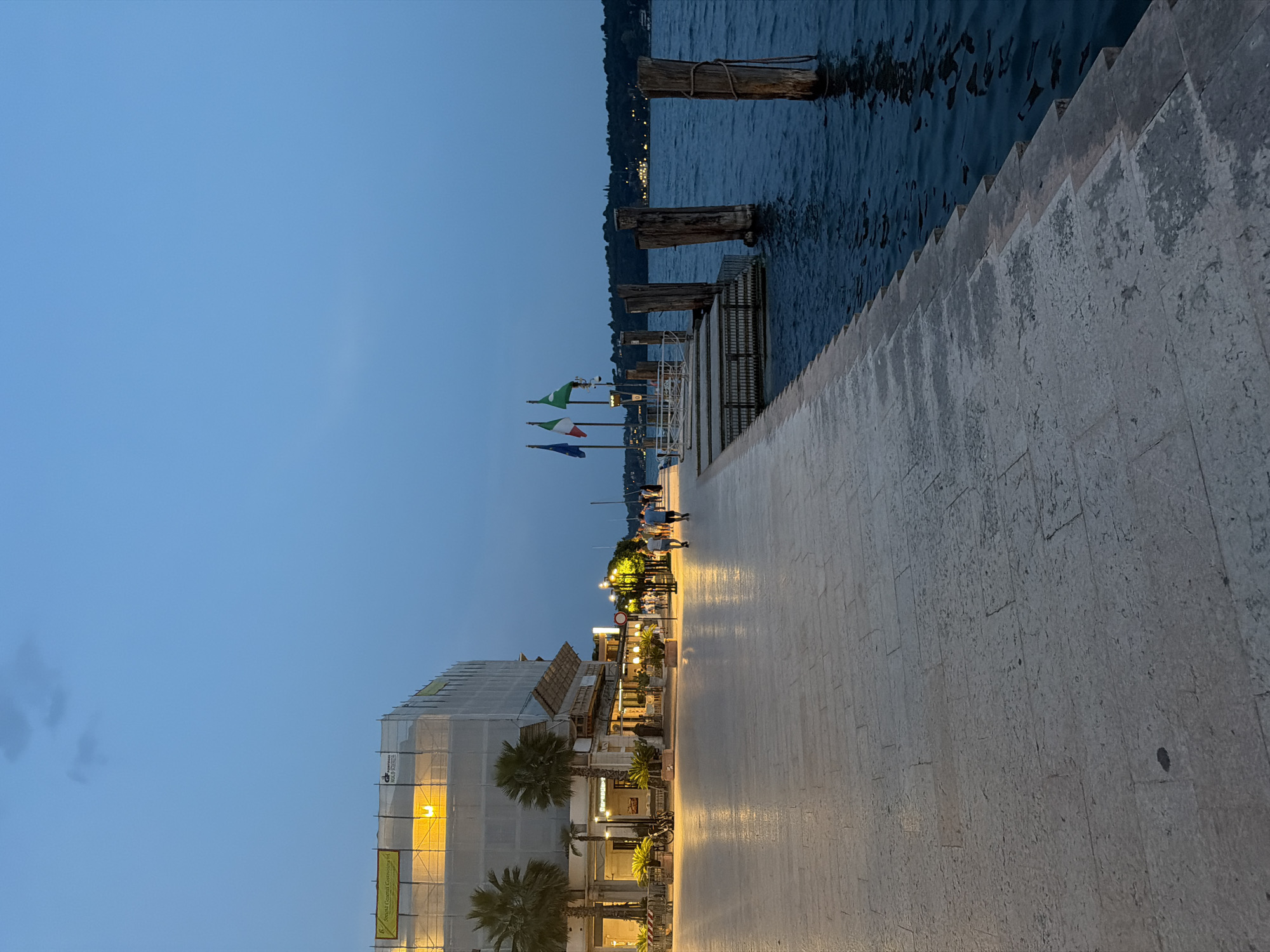 The image depicts a waterfront promenade at dusk in Salò, Italy. The scene is characterized by a wide, paved walkway with a stone surface, illuminated by warm streetlights that cast long shadows. To the left, a building is under scaffolding, with brightly lit windows visible through the white sheeting. Palm trees and potted plants line the walkway in front of restaurants and shops. Several people are walking along the promenade.
To the right of the walkway, the dark blue water of Lake Garda laps against the shore. A set of wooden pilings, weathered and partially submerged, mark the edge of the water. A small dock with tiered steps leads down to the water, adorned with three flags: one appears to be the Italian flag, another green with a white emblem, and a third with blue and yellow. In the distance, across the lake, the lights of buildings and a tree-lined shore are visible. The sky is a deep twilight blue with a few faint clouds.