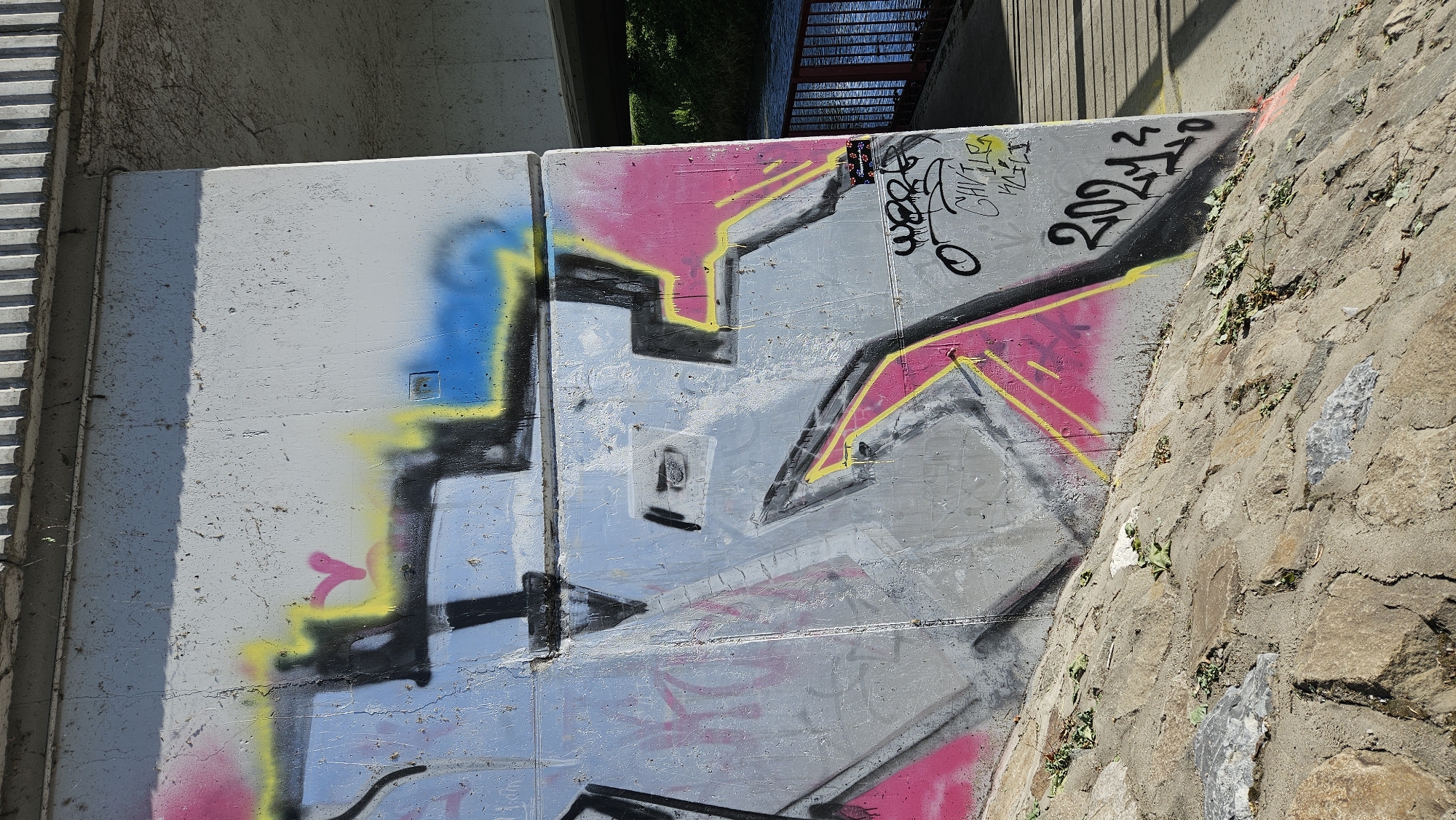 This image shows graffiti on a concrete support structure, likely part of an overpass or bridge, near a body of water. The graffiti includes abstract shapes and lines in black, yellow, and pink spray paint. There is also text that reads "2021". A small sticker with multiple colorful squares is affixed to the concrete. In the background, a body of water with a railing can be seen, along with some greenery. The concrete structure is positioned on a rocky embankment. Based on the provided information, this scene is located in Prešov, Slovakia.