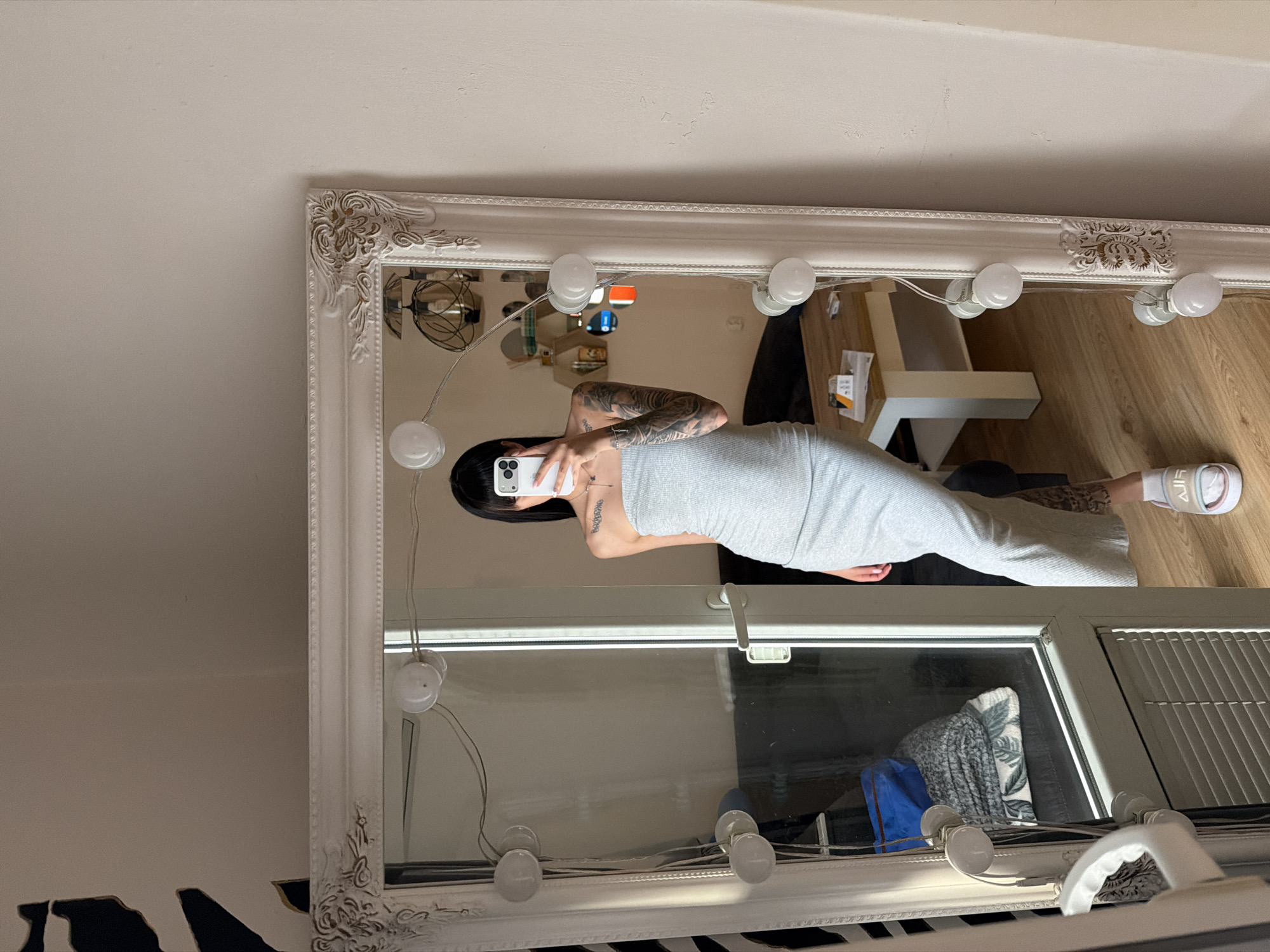 A young woman with dark hair, wearing a light grey strapless midi dress and white slides, stands in front of a large ornate mirror with lights around its edge. She is holding a smartphone in her right hand, taking a selfie. Her arms and shoulders are covered in tattoos. The mirror reflects a room with a wooden floor, a white coffee table, and some shelving with decorative items. The scene appears to be a domestic setting, likely a bedroom or dressing area. The overall impression is one of personal style and self-expression. There is no indication of the city of Plovdiv, Bulgaria.