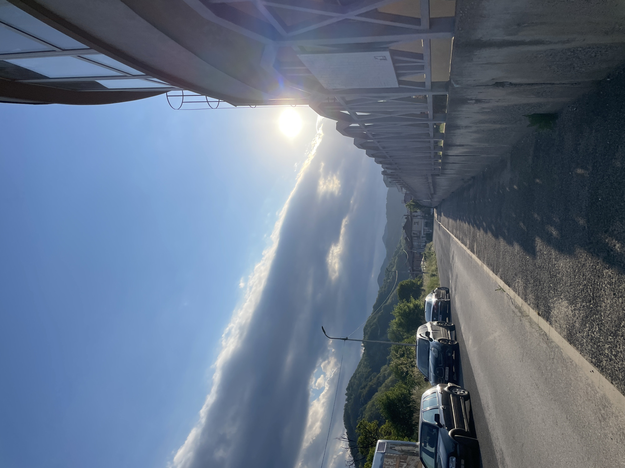 The image shows a street in Zlatograd, Bulgaria, lined with parked cars. The perspective is looking down the road with a view of the surrounding mountains and a dramatic cloud formation in the sky. A building overhangs the left side of the frame, casting a shadow. The sun is visible in the distance, contributing to a bright and high-contrast scene.