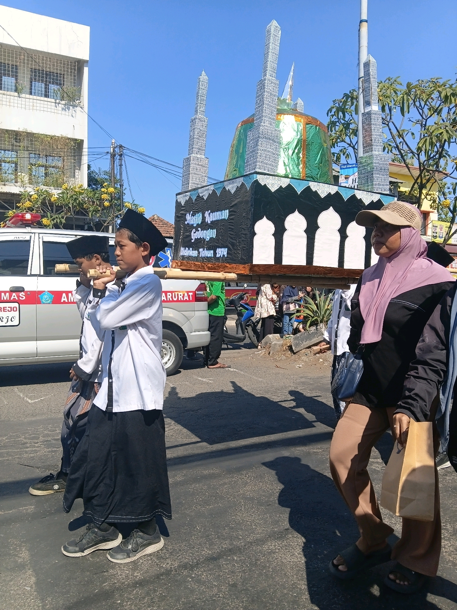 In Wadungasri, Indonesia, on a bright, sunny day, a procession is underway. Two young boys, dressed in white long-sleeved shirts and black peci hats, carry a large, decorated float on their shoulders using wooden poles. The float resembles a mosque, complete with a green dome, minarets, and a banner that reads "Masjid Kauman Gedongan Didirikan Tahun 1974." Behind the boys, a woman wearing a pink hijab and a tan baseball cap walks, holding a brown paper bag. A white ambulance with red and blue markings is visible to the left of the procession. The scene appears to be taking place on a paved road, with buildings, trees, and a clear blue sky in the background.