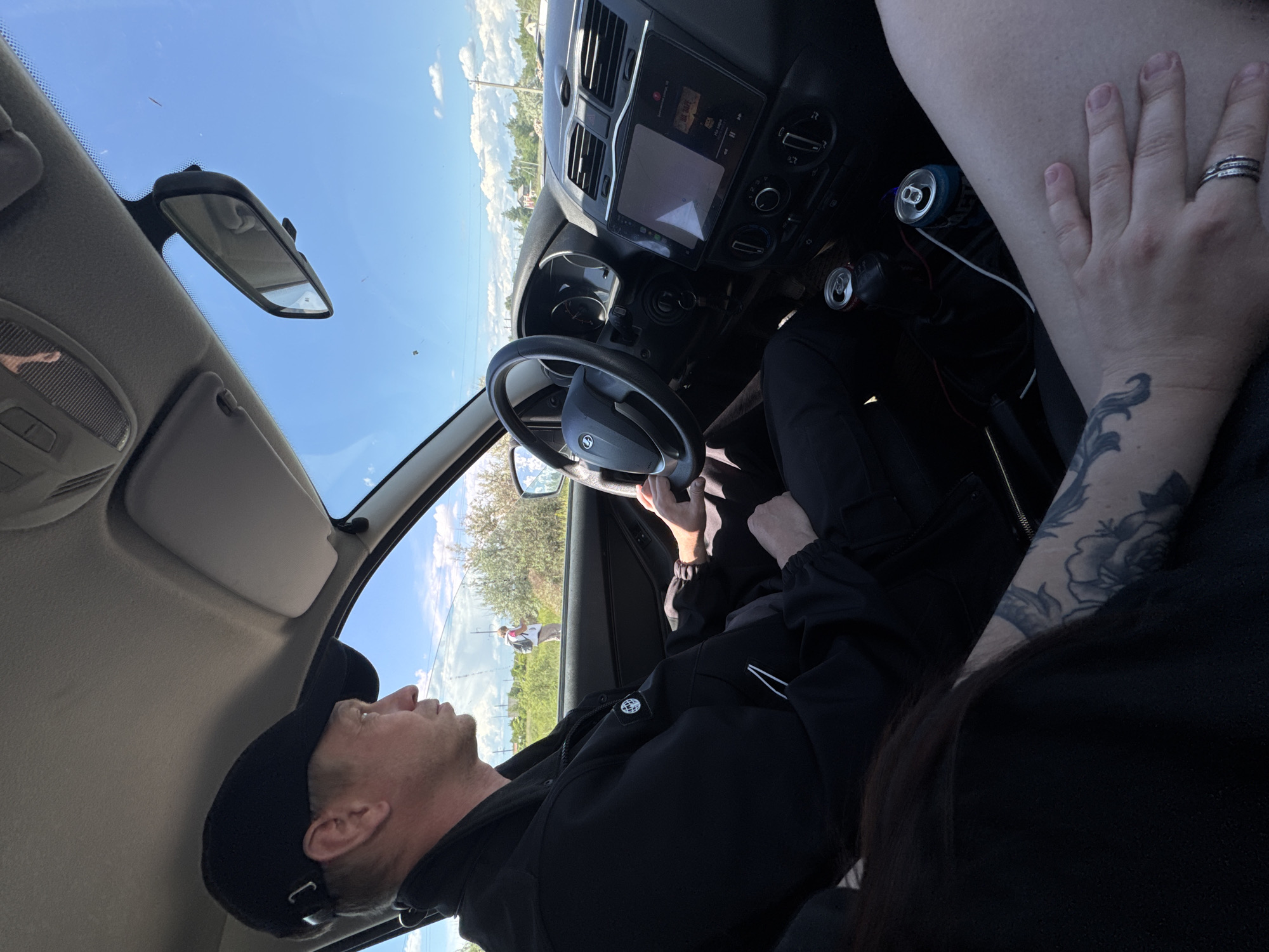The interior of a car is depicted, with a man wearing a black cap and jacket visible in the driver's seat, his hands on the steering wheel. A woman's hands and tattooed arm are visible in the passenger seat. The car's dashboard is visible, along with the rearview mirror. Outside the window, there is a view of a landscape with trees, grass, and utility poles, and a person walking. The location is Unknown, Unknown.