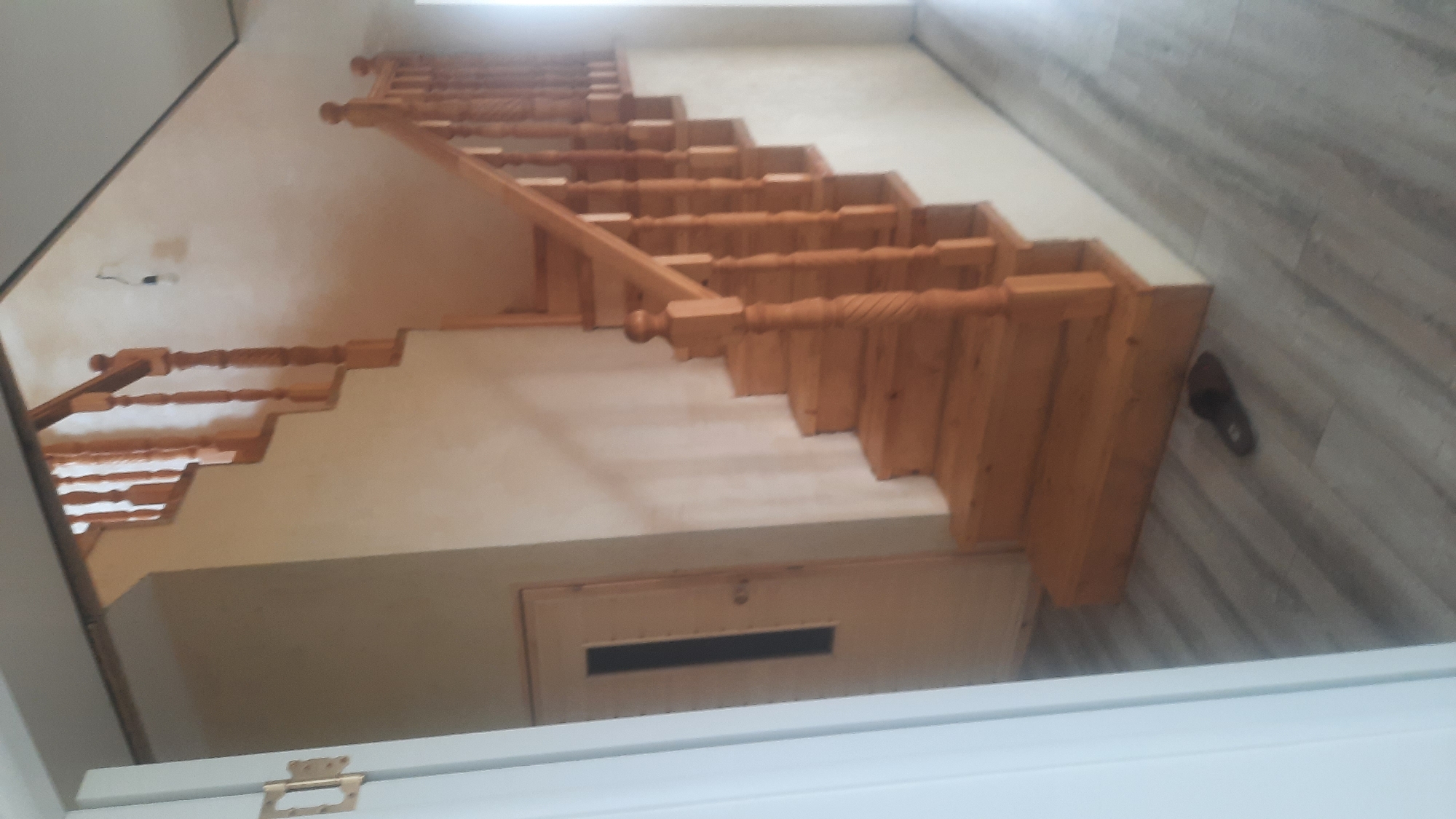 A wooden staircase is visible in a residential interior, likely in the city of Қаражар, Kazakhstan. The staircase, constructed from light-colored wood, features carved balusters and a wooden handrail. The walls surrounding the staircase are painted a neutral, light beige color. To the left of the staircase, a door with a narrow, dark rectangular window is present. The floor is covered with light gray laminate flooring with a wood grain pattern. A single dark slipper is positioned on the floor near the bottom of the staircase. The lighting suggests it is daytime, with natural light entering from an unseen source, casting shadows on the stairs and floor.