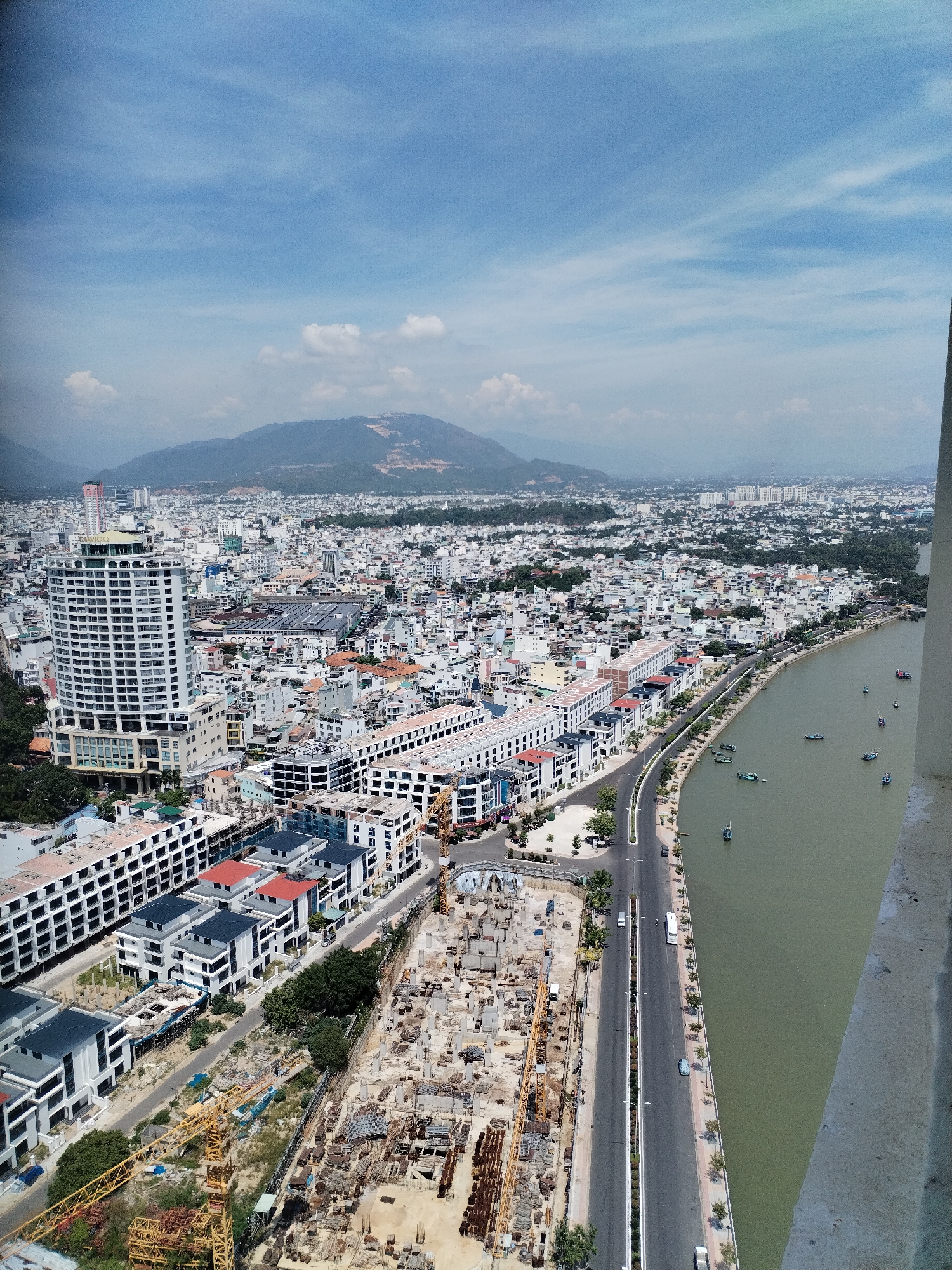 This is an aerial, high-angle shot of Nha Trang Ward, Vietnam, captured on a bright, slightly hazy day. The scene is dominated by a sprawling cityscape with a prominent river or bay curving through it. The foreground on the right is a concrete balcony or edge of a building, indicating the vantage point.
The city itself is a dense collection of buildings, varying in height and architectural style, with many modern, white structures and some older, lower-rise buildings. A large, distinctive white, circular high-rise building is a focal point in the mid-left of the frame, labeled with "KHANHHOA". Adjacent to it, and stretching along the river, are newer, multi-story residential or commercial buildings, many with white facades and red or dark-tiled roofs.
A significant area in the mid-ground is a large construction site, characterized by exposed concrete foundations, rebar, and multiple yellow construction cranes. A road runs parallel to the river, lined with trees and some vehicles, including cars and a white van.
The river itself is a murky green color and several small boats, likely fishing or transport vessels, are scattered on its surface. Beyond the city, a range of blue-toned mountains forms the backdrop, with some evidence of development or construction on their slopes. The sky is a bright blue with scattered white clouds. The overall impression is of a rapidly developing coastal city.