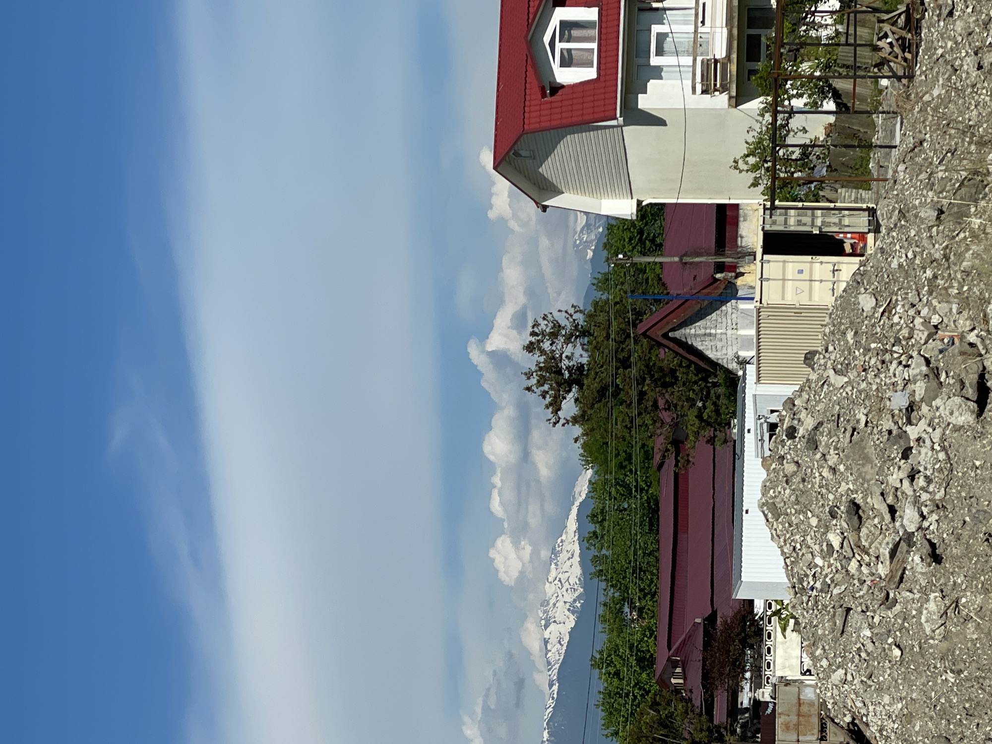A daytime outdoor scene in Unknown, Unknown, depicts a landscape composed of construction remnants, residential structures, dense vegetation, and distant snow-capped mountains under a partly cloudy sky. In the immediate foreground, a substantial pile of grey rubble, consisting of concrete fragments, stones, and earth, occupies the lower central and left portions of the frame. To the right of the rubble, sparse dry grass and a dark metal frame structure are visible.
Behind the rubble, a light-colored shipping container with an open door reveals an orange traffic cone inside. To its left, a white corrugated metal shed and a rusty metal gate are partially obscured by the debris.
The midground features several residential buildings interspersed with lush green trees. On the far right, a two-story house with a prominent red tiled gable roof and white siding features a white-framed window and a balcony on its upper level. Towards the center-left, multiple buildings with dark red tiled roofs are partially visible amongst the trees; one exhibits an A-frame roof. Overhead utility lines stretch across the midground above the tree line.
In the background, a range of snow-capped mountains spans the horizon beneath a bright blue sky. Scattered white cumulus clouds populate the sky, with clearer blue above. No human figures or discernible activities are present.