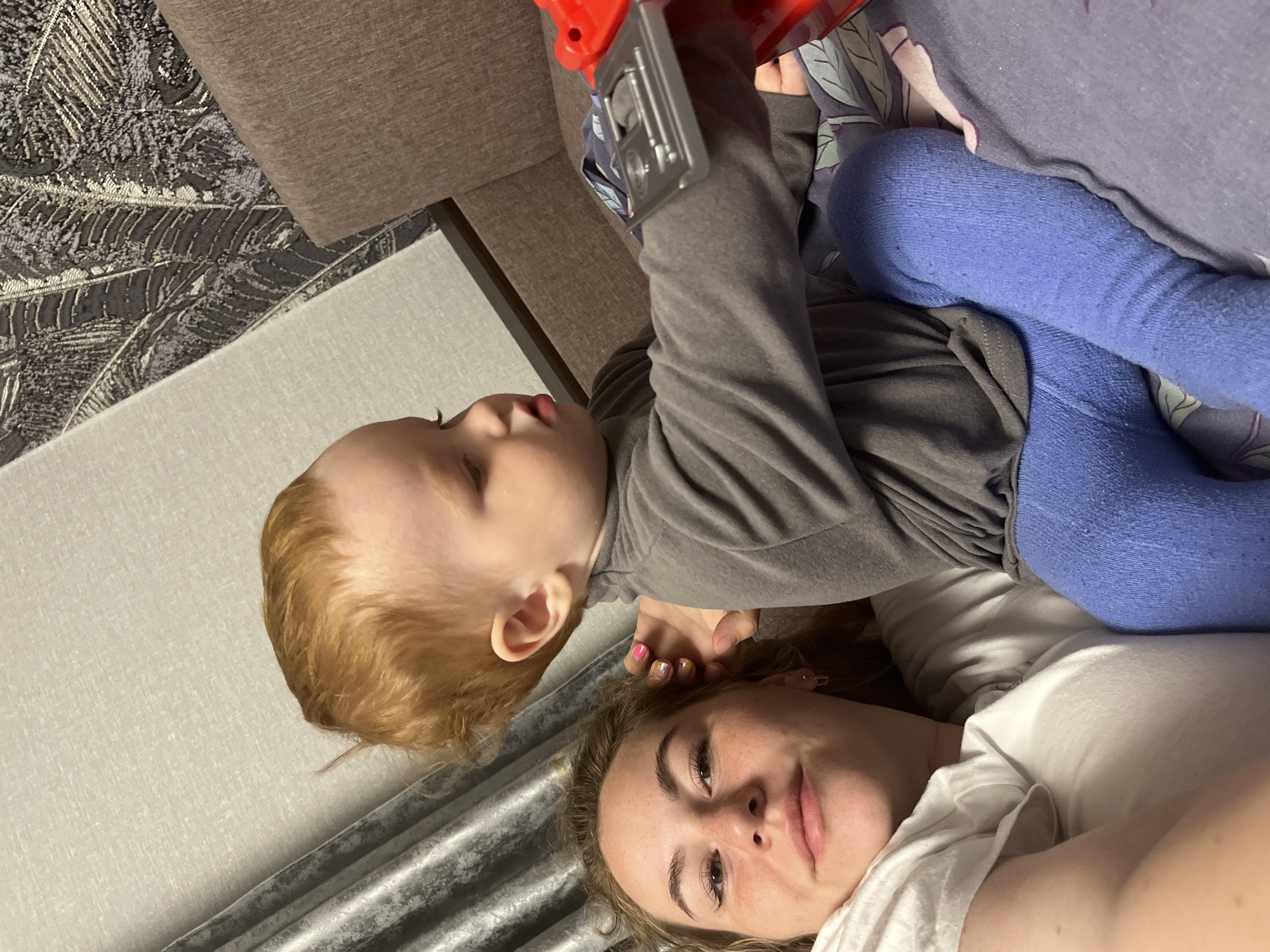 A woman and a young child are lying on a bed or sofa, likely indoors. The woman is wearing a white t-shirt, and the child is in a gray long-sleeved shirt and blue leggings. The child is holding a red and gray toy. The location may be Chaikovsky, Russia. The setting appears to be a domestic environment.