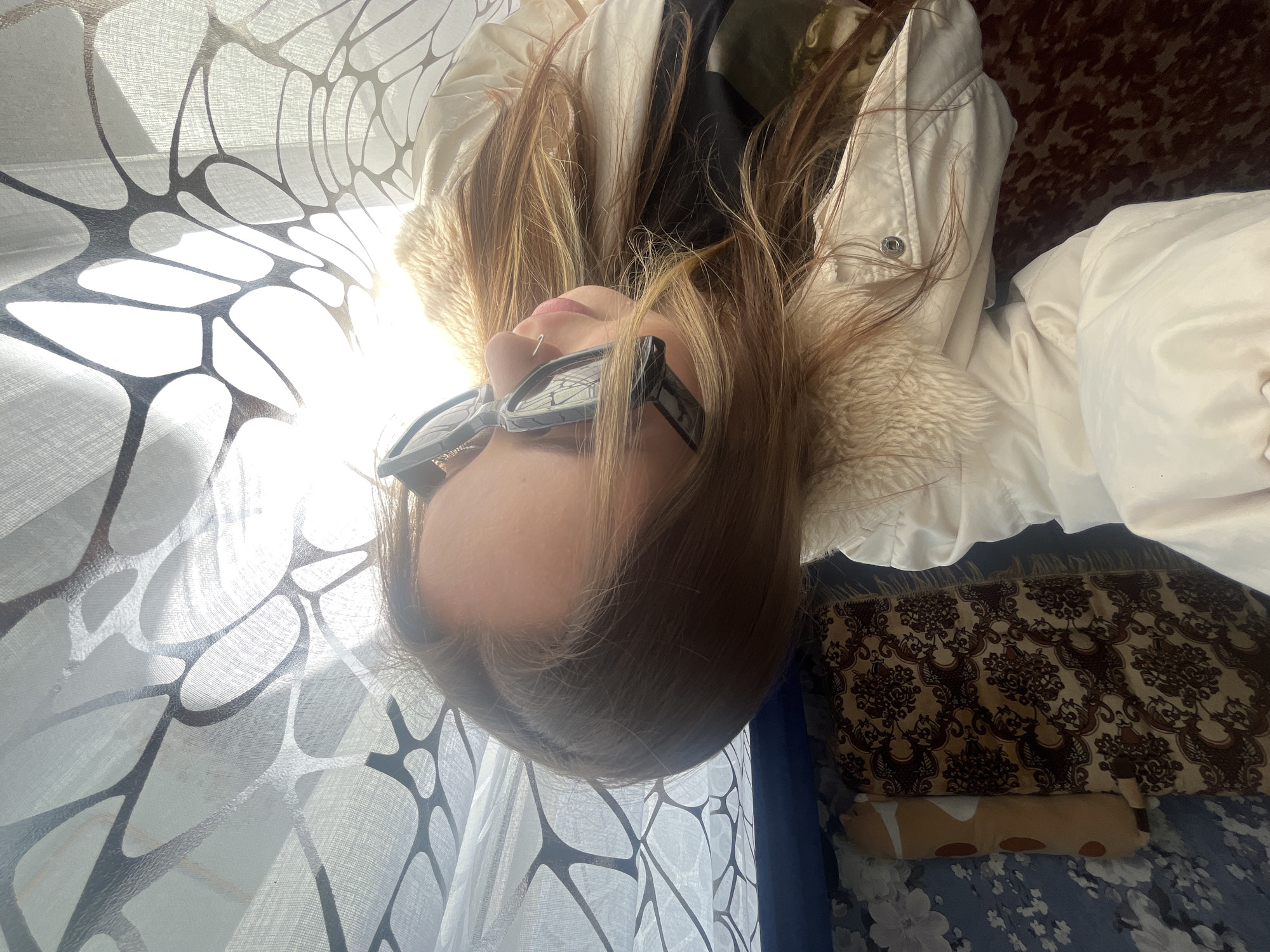 A person wearing sunglasses lies in front of a window covered by a white curtain with a dark, organic pattern. The person is wearing a white jacket with a fur-like collar. A pillow with a brown and beige floral pattern is partially visible. The bed has a blue and white floral pattern. Novopokrovskaya, Russia, is not discernable from this image.