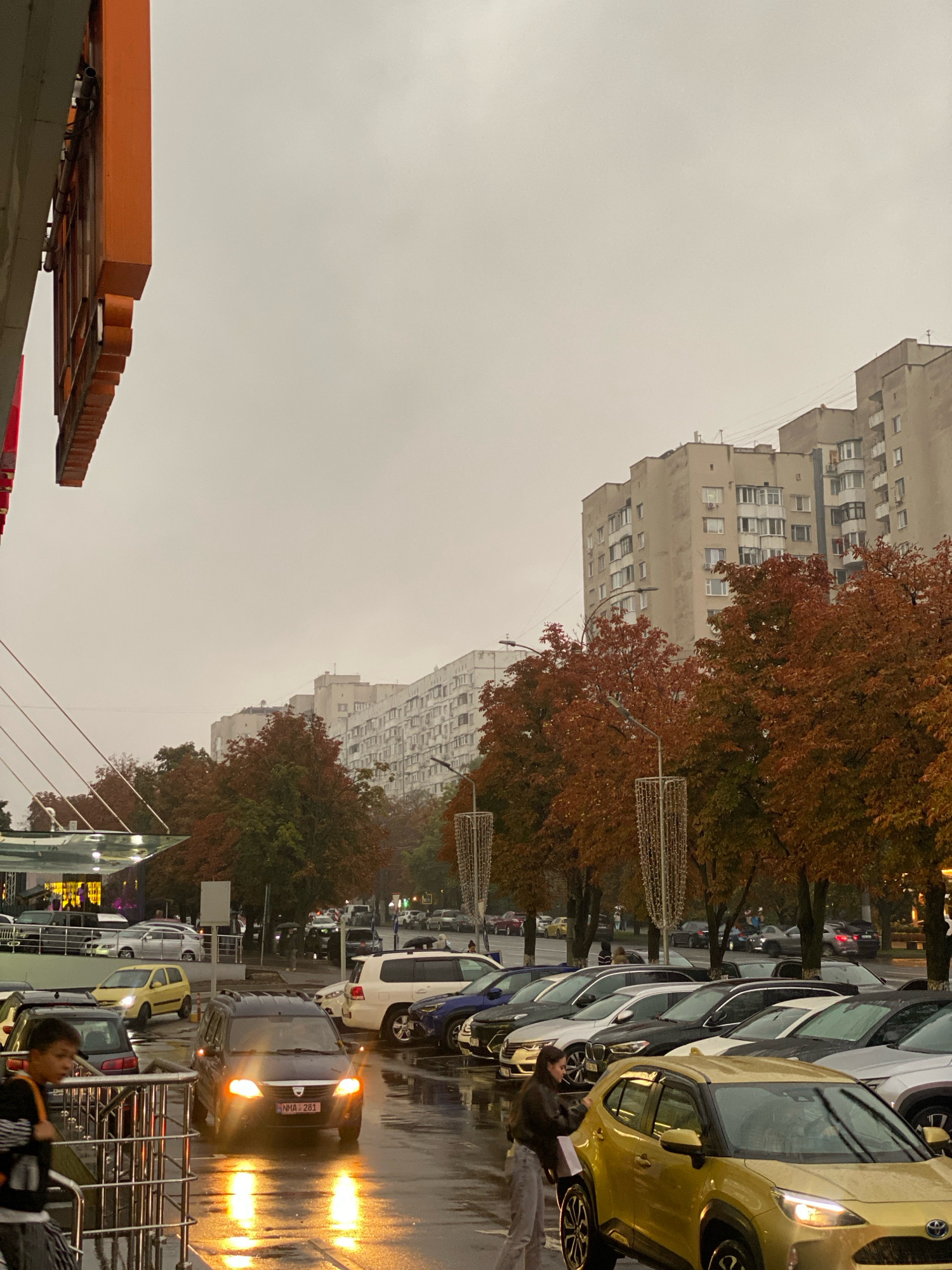 The image depicts a street scene in Chișinău, Moldova, on an overcast day. The foreground features a parked yellow car and a dark minivan with its headlights on, reflecting on the wet pavement, indicating recent rain. A person, possibly a teenager, is visible to the left, leaning on a railing. In the middle ground, several cars are parked in rows, and people are seen walking. The background consists of apartment buildings and trees with autumn foliage. The overall atmosphere suggests a typical urban environment during the fall season.
