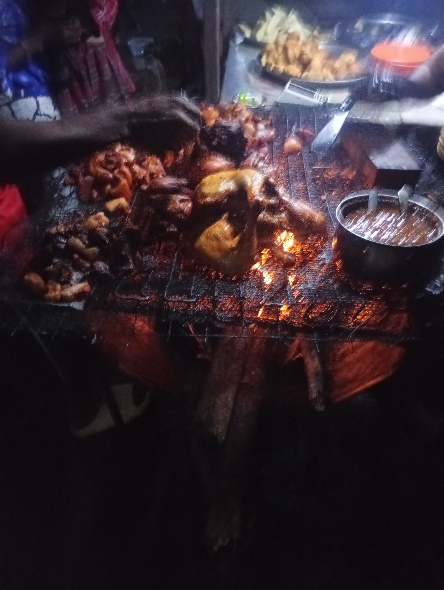 An outdoor nighttime scene depicts a close-up of various pieces of food, likely meat, being grilled over an open fire. The food is arranged on a metal grate, with visible flames and embers below. To the right, a metal bowl filled with a dark, viscous liquid, possibly a sauce or dip, sits near the edge of the grate. In the background, blurred figures and objects suggest a street food vendor setting. The overall lighting is dim, with the fire providing the primary illumination. The location is described as Unknown, Unknown.