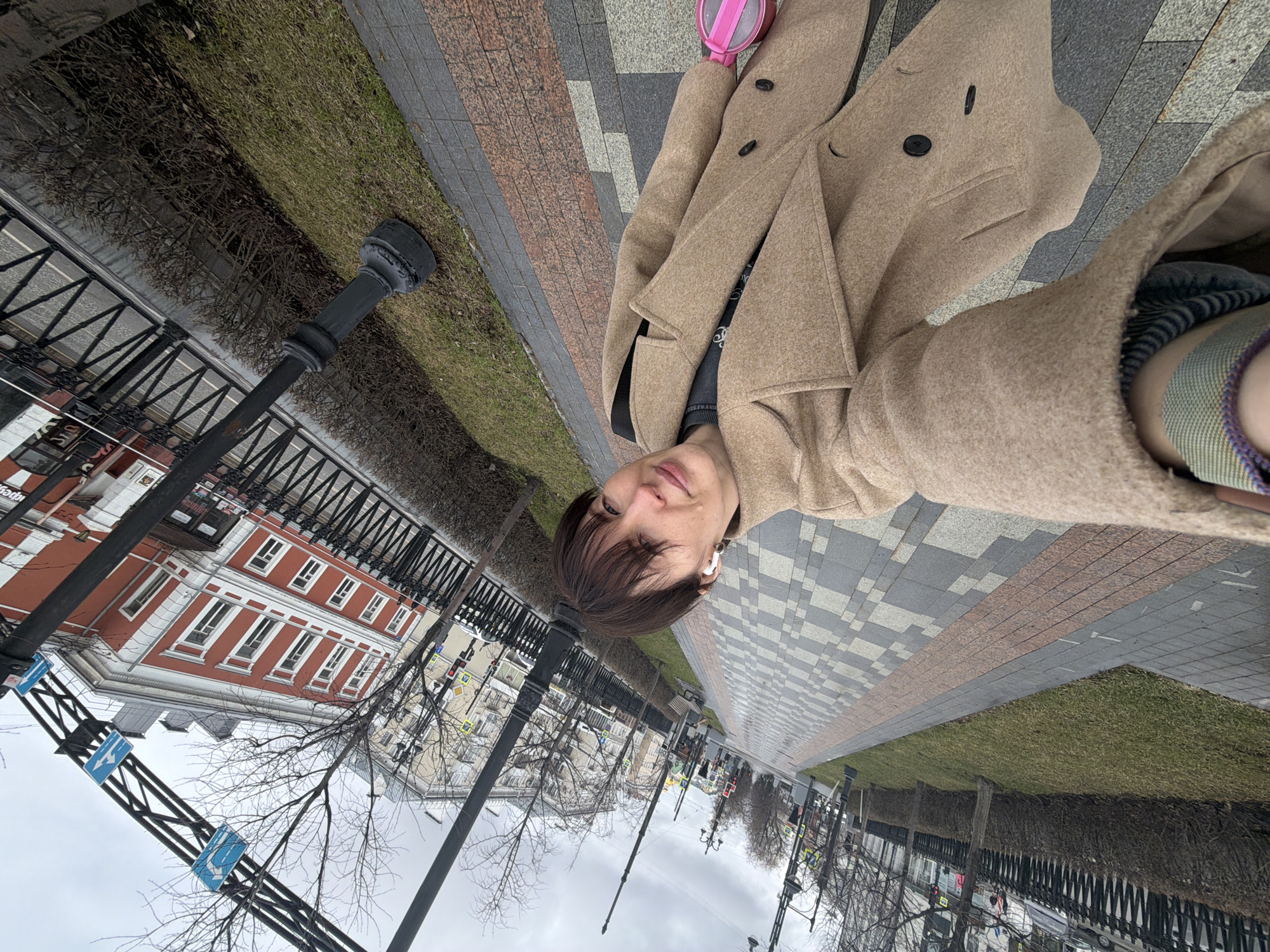 A selfie taken on a city street in Perm, Russia. The individual is wearing a tan coat and a white wireless earbud. Behind them is a sidewalk with patterned brickwork, a strip of grass, and a black wrought-iron fence. In the background are buildings with red and white facades, street lights, bare trees, and a cloudy sky.