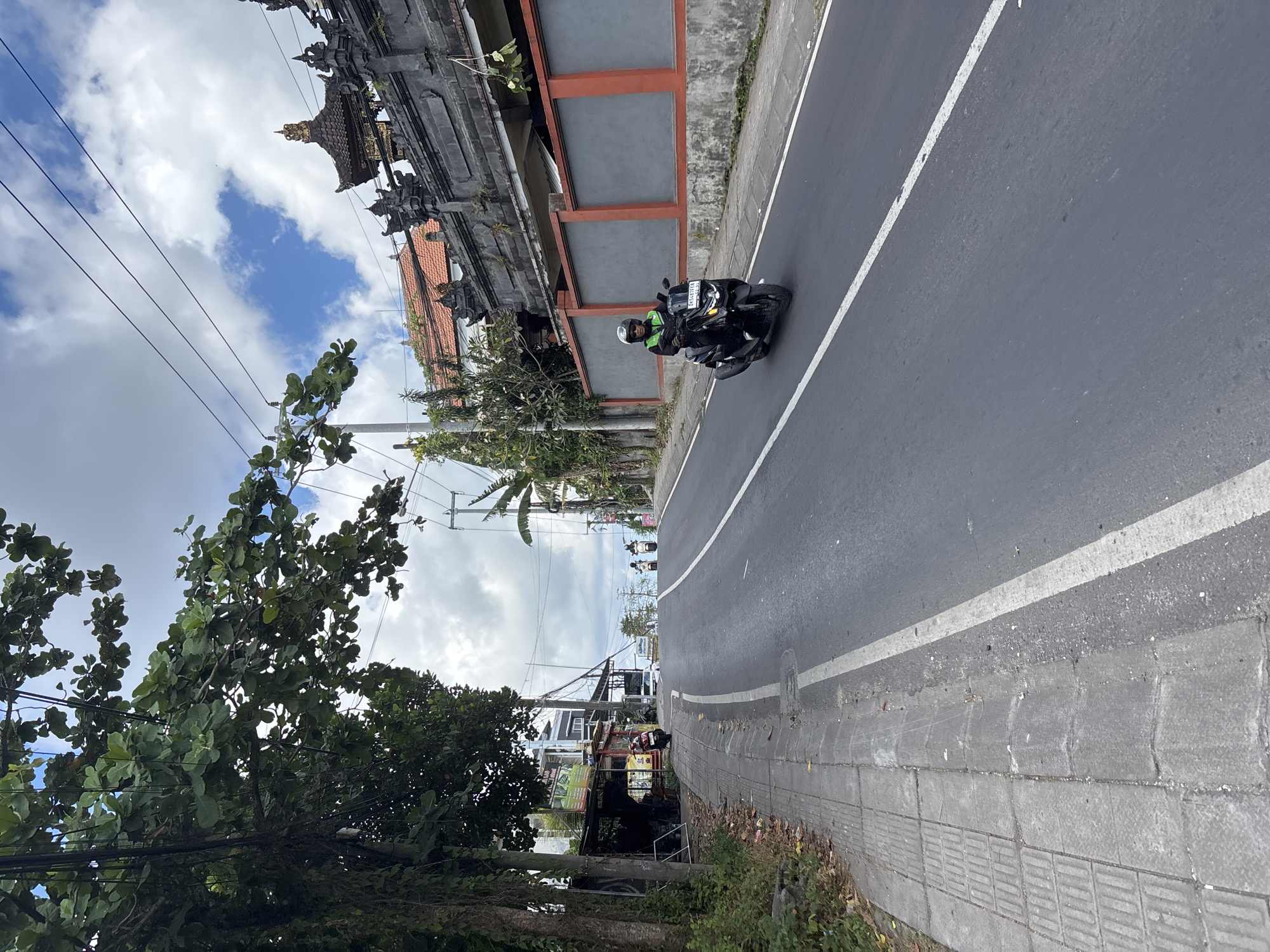 This media file captures a daytime street scene in Kuta Selatan, Indonesia, depicting a road with adjacent buildings and lush vegetation under a partially cloudy sky. The image appears to be rotated 90 degrees counter-clockwise.
In the foreground and mid-ground, a dark-colored motorcycle is visible on the asphalt road, traveling away from the viewer. A person wearing a helmet and a bright green or yellow top is riding the motorcycle. The license plate "DD 6454 ED 07-29" is clearly visible on the front of the motorcycle, indicating a registration from the Denpasar area of Bali and an expiration date of July 2029. The road itself features a solid white line marking the edge of the lane or shoulder.
To the left of the road, there is a concrete sidewalk, some sections of which have tactile paving patterns. Beyond the sidewalk, traditional Balinese architecture is prominent, featuring ornate, dark-colored buildings with intricate carvings and multi-tiered roofs, interspersed with red-tiled roofs and grey walls with red trim. Further down the road, more commercial or residential structures are visible, though indistinct.
The right side of the scene is dominated by dense, green tropical foliage, including a large leafy tree in the immediate foreground and palm trees further back. Overhead, numerous black power lines crisscross the bright blue sky, which is dotted with white clouds. The overall impression is one of a typical bustling street in a tropical Indonesian setting.
