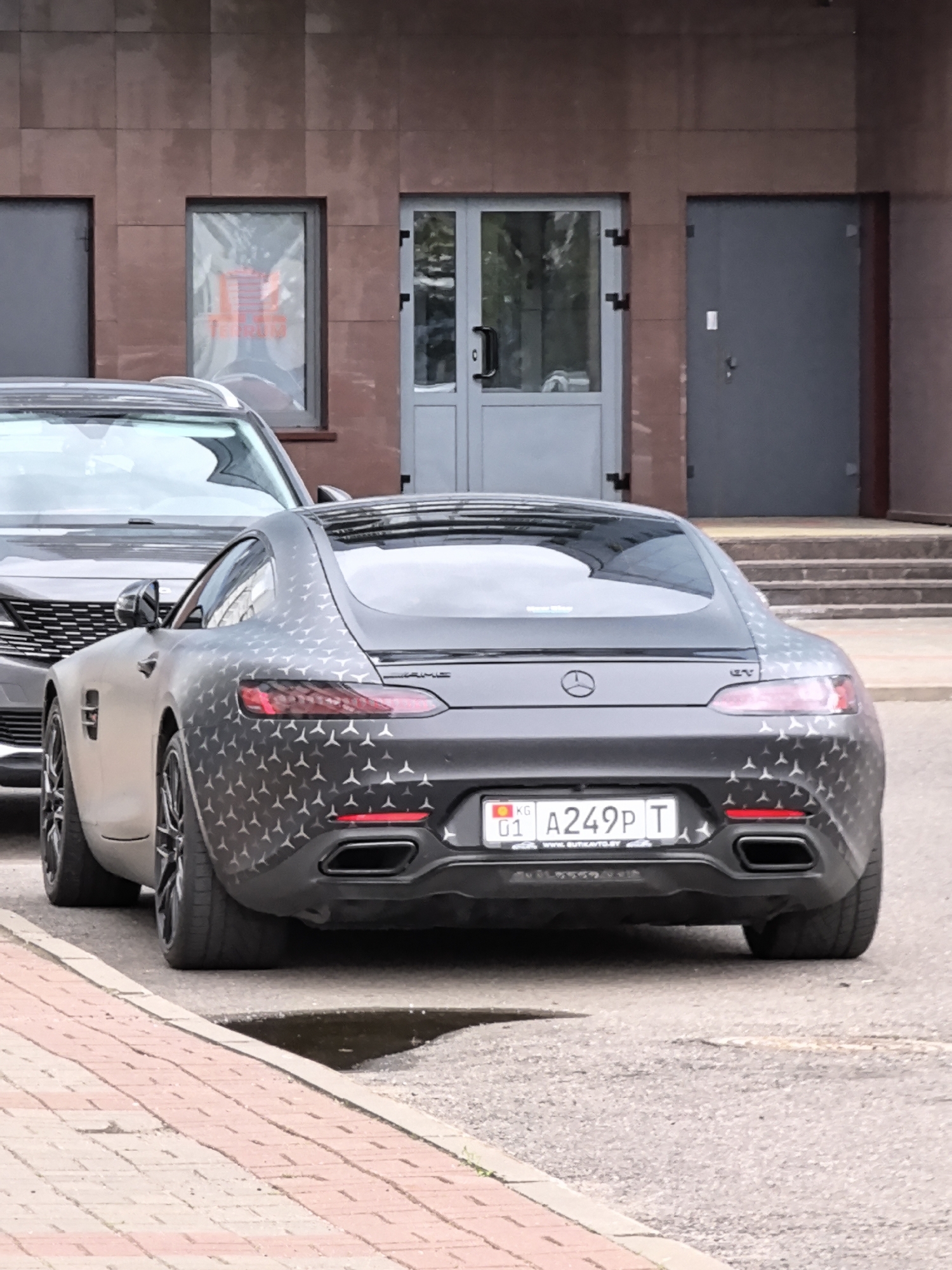 A rear-view, eye-level shot shows a black Mercedes-Benz AMG GT car parked on an asphalt street next to a brick sidewalk. The car has a custom wrap with a repeating pattern of small silver star emblems. The license plate, reading "KG 01 A249P T", is visible. A portion of a gray SUV is visible to the left of the AMG GT. In the background, a building with large glass doors and windows is present. The ground is wet in one area, suggesting recent rain. The lighting suggests it is daytime.