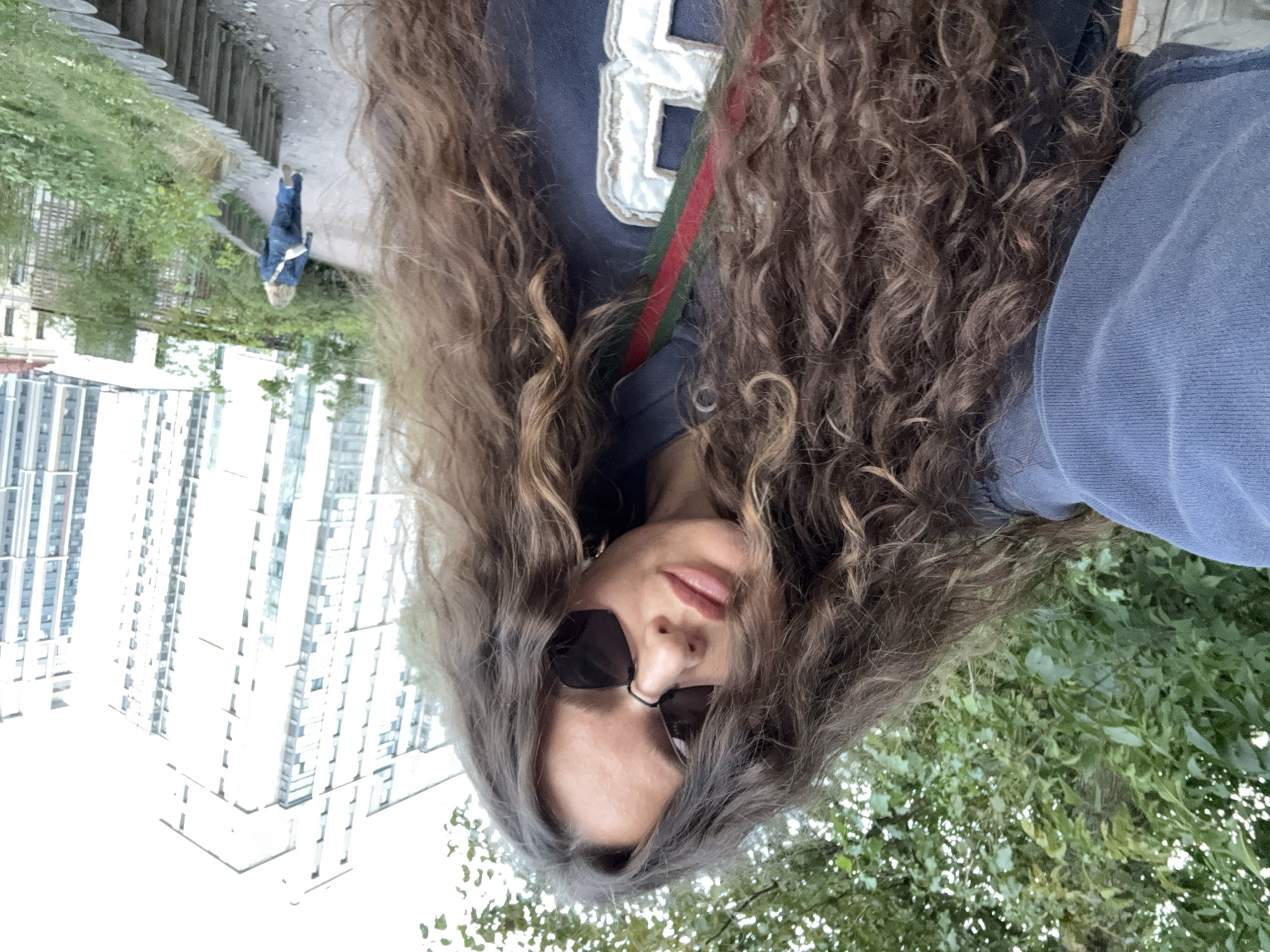 A woman with long, curly brown hair and sunglasses is taking a selfie outdoors. She is wearing a blue hoodie with a green and red stripe and a white embroidered number. In the background, there are tall buildings and a walkway lined with wooden posts. Another person in a denim outfit is walking away on the walkway. The scene appears to be a park or urban green space.