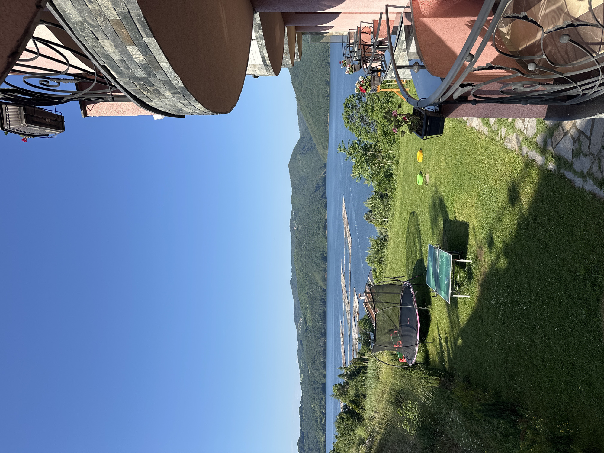 The image depicts a scenic view of a lake, likely from a balcony or elevated vantage point, in Glavatartsi, Bulgaria. The foreground is dominated by a balcony with a pink facade and a dark metal railing. A potted plant with pink flowers is visible on the balcony. Below the balcony, a green lawn features a trampoline with a black net and a green and white ping-pong table. Two colorful rings, one green and one yellow, are placed on the grass. Beyond the lawn, the expansive blue water of the lake stretches towards a backdrop of lush green, tree-covered hills and mountains under a clear blue sky. In the middle of the lake, a series of floating structures, possibly fish farms or rafts, are arranged in rows. The overall scene suggests a recreational outdoor setting in a picturesque natural environment.
