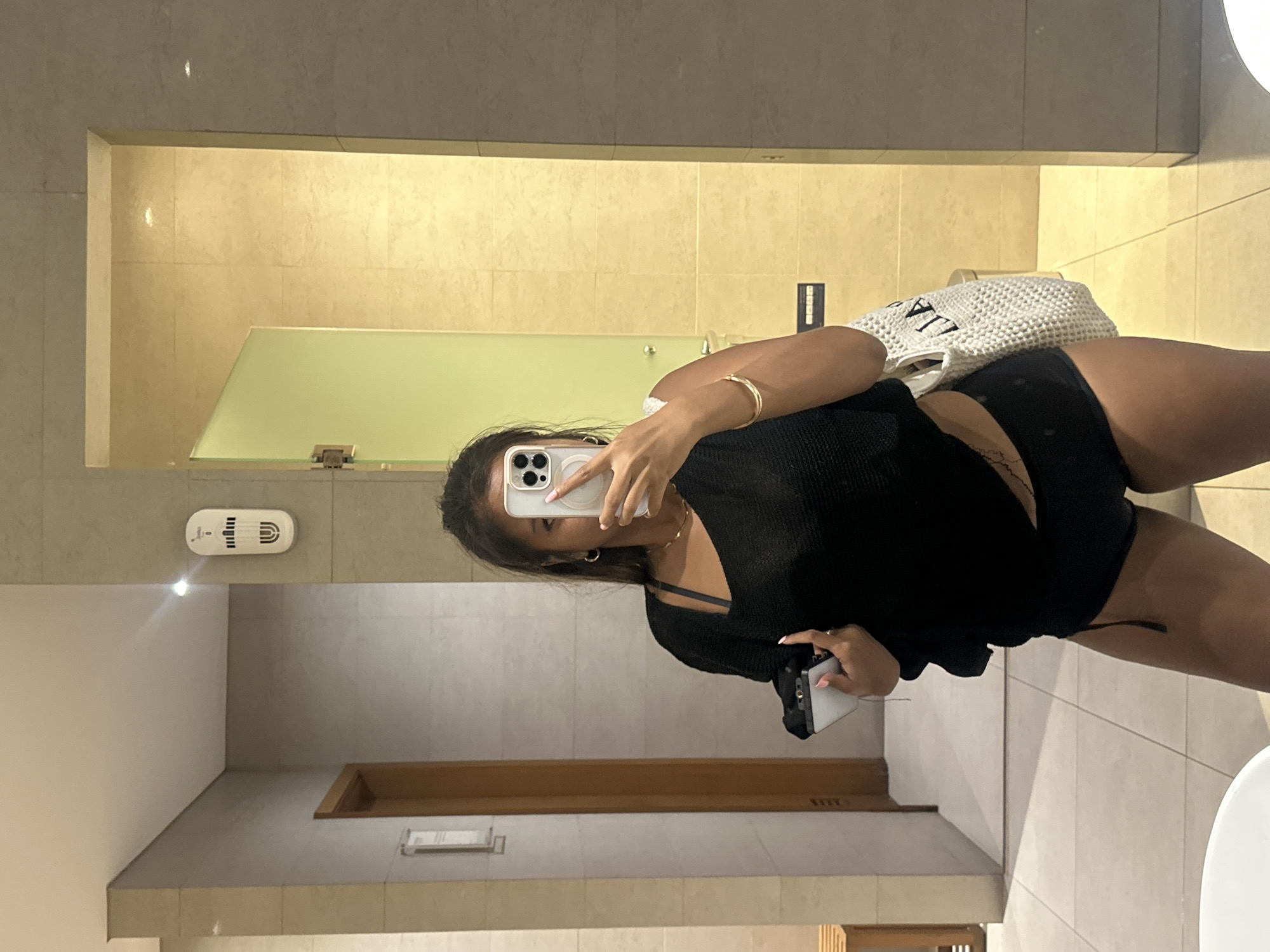 A woman is taking a selfie in a bathroom. She is wearing a black top and black shorts. The bathroom has a tile floor and tile walls. There is a white bag hanging from her shoulder. There is a door in the background. A smoke detector is visible on the wall. The location is Central Jakarta, Indonesia.