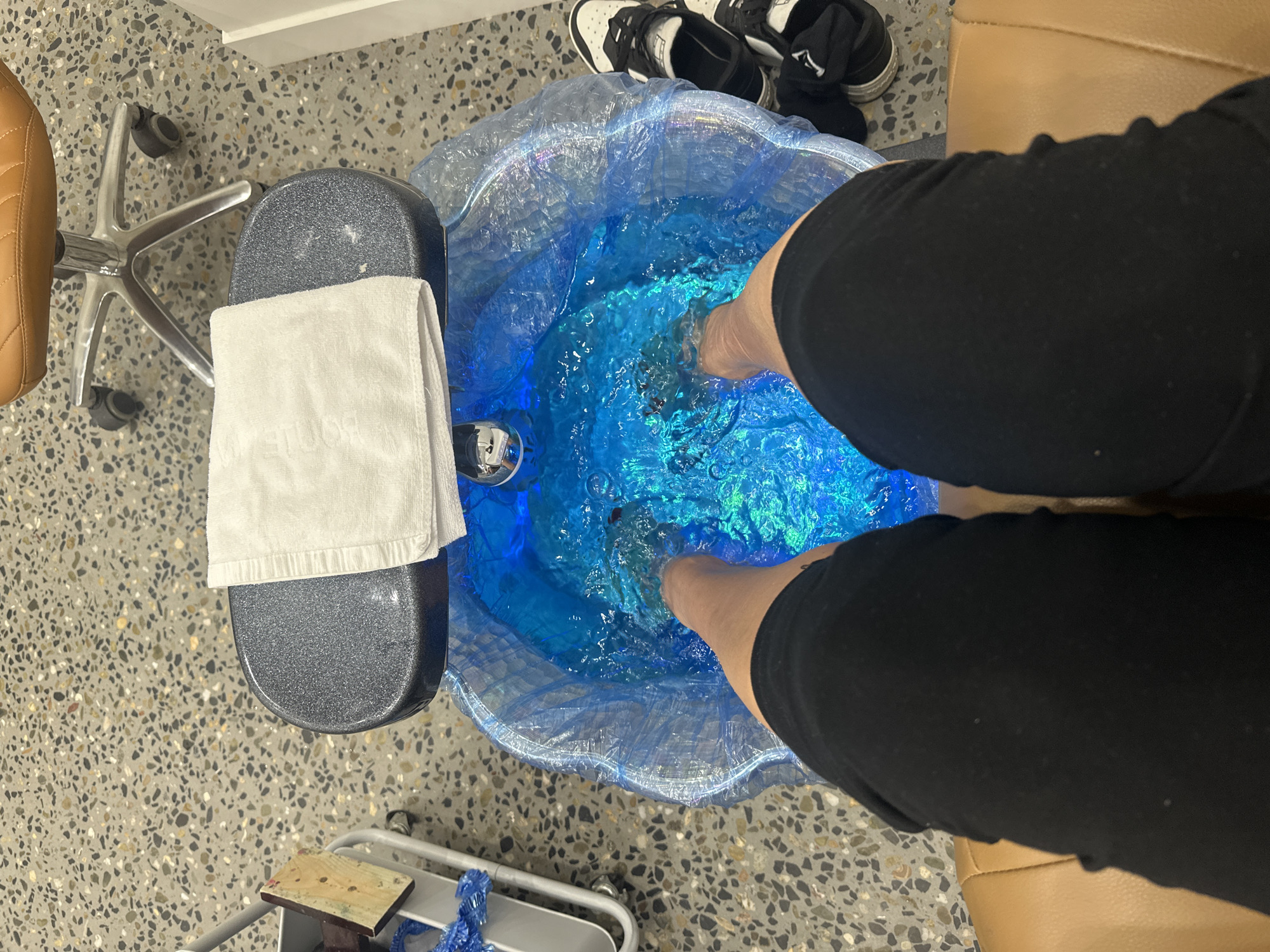 The image depicts a person's legs and feet submerged in a pedicure basin filled with luminous blue water. The basin is lined with a clear plastic bag. A white towel is draped over the edge of the basin. The person is seated in a tan pedicure chair, and their legs are covered by black shorts. In the background, to the right, a pair of black and white sneakers are visible. To the left, a metal trolley with a wooden piece and some blue plastic material is present. The floor has a speckled, terrazzo-like pattern. The scene appears to be in a nail salon, possibly located in Coffs Harbour, Australia, given the provided context.