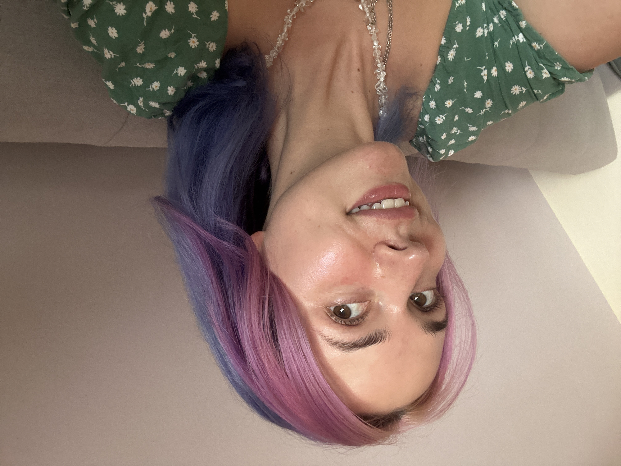A woman is shown in an upside-down close-up shot. She has purple and pink dyed hair and brown eyes. She is wearing a green dress with small white floral patterns and a crystal necklace. She is smiling slightly and lying on a grey cushion or sofa. The background appears to be a plain, light-colored wall. The location is not discernible from the image.