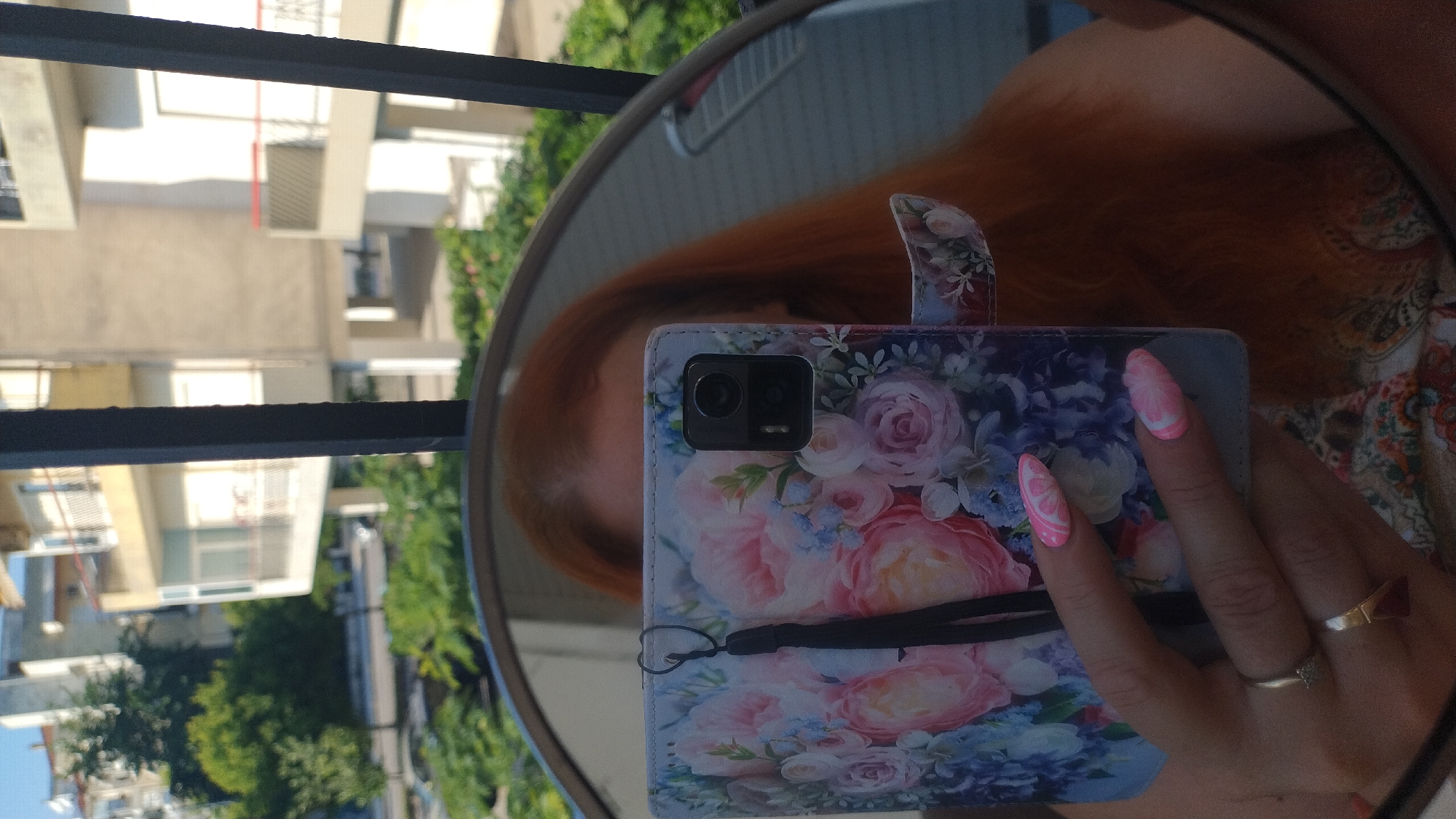 A woman with auburn hair is reflected in a round mirror, holding a floral patterned phone case. Her nails are painted pink with white designs. The background of the reflection shows buildings and trees, suggesting an urban environment.