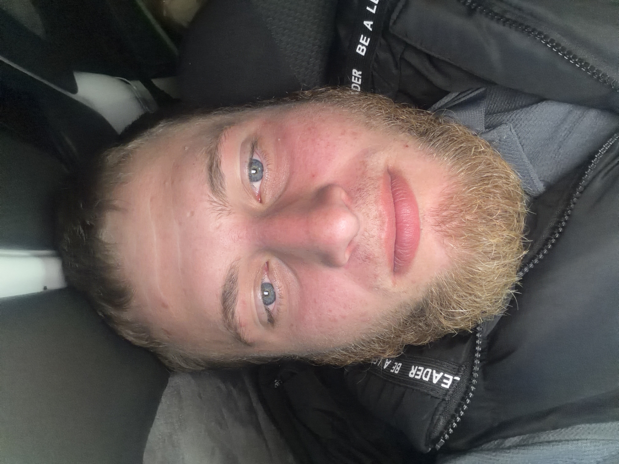A close-up, rotated selfie of a young Caucasian male with light brown hair and a light brown beard is shown. He has blue eyes, fair skin with some redness, and pink lips. He is wearing a dark jacket with a zipper and the words "LEADER BE A L" visible on a white label, and a grey collared shirt underneath. The background appears to be dark and possibly indoors, with a greyish area on the upper left and a black area on the right. The image is oriented such that the man's head is towards the bottom of the frame. There is no discernible indication of the city of Sculeni, Moldova within the image itself.