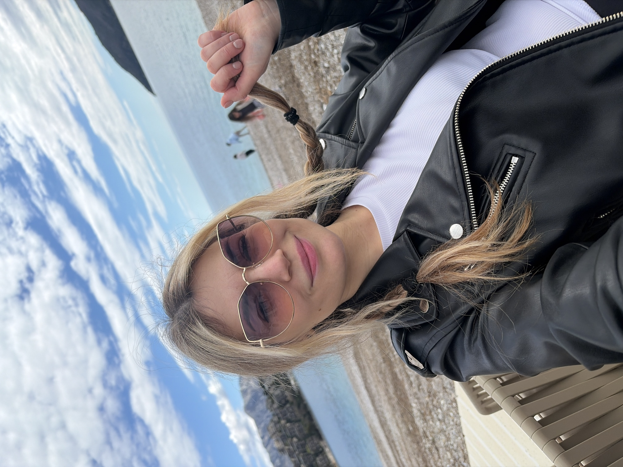 A woman poses for a photo on a beach in Budva, Montenegro. She has blonde hair styled in a braid and wears sunglasses, a white shirt, and a black leather jacket. The background includes a pebble beach, the sea, a cloudy sky, and a rocky cliff face. Several other people are visible in the distance on the beach. She is seated in a beige plastic chair.