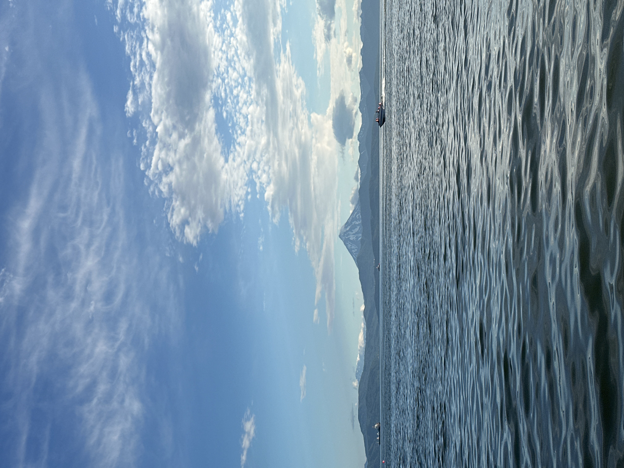 A body of water, likely a bay or harbor, in Petropavlovsk-Kamchatsky, Russia, under a partly cloudy sky. The water has moderate ripples, reflecting the sky and surrounding landscape. In the distance, dark, rounded mountains form the backdrop, with a prominent, snow-capped conical volcano dominating the center. On the water, two small vessels are visible: a larger ship with a white superstructure and dark hull is to the left, and a speed boat with several occupants is moving from left to right on the right side of the frame. The sky transitions from a clear blue at the top to a band of white, wispy clouds, and then to larger, puffy cumulus clouds closer to the horizon.