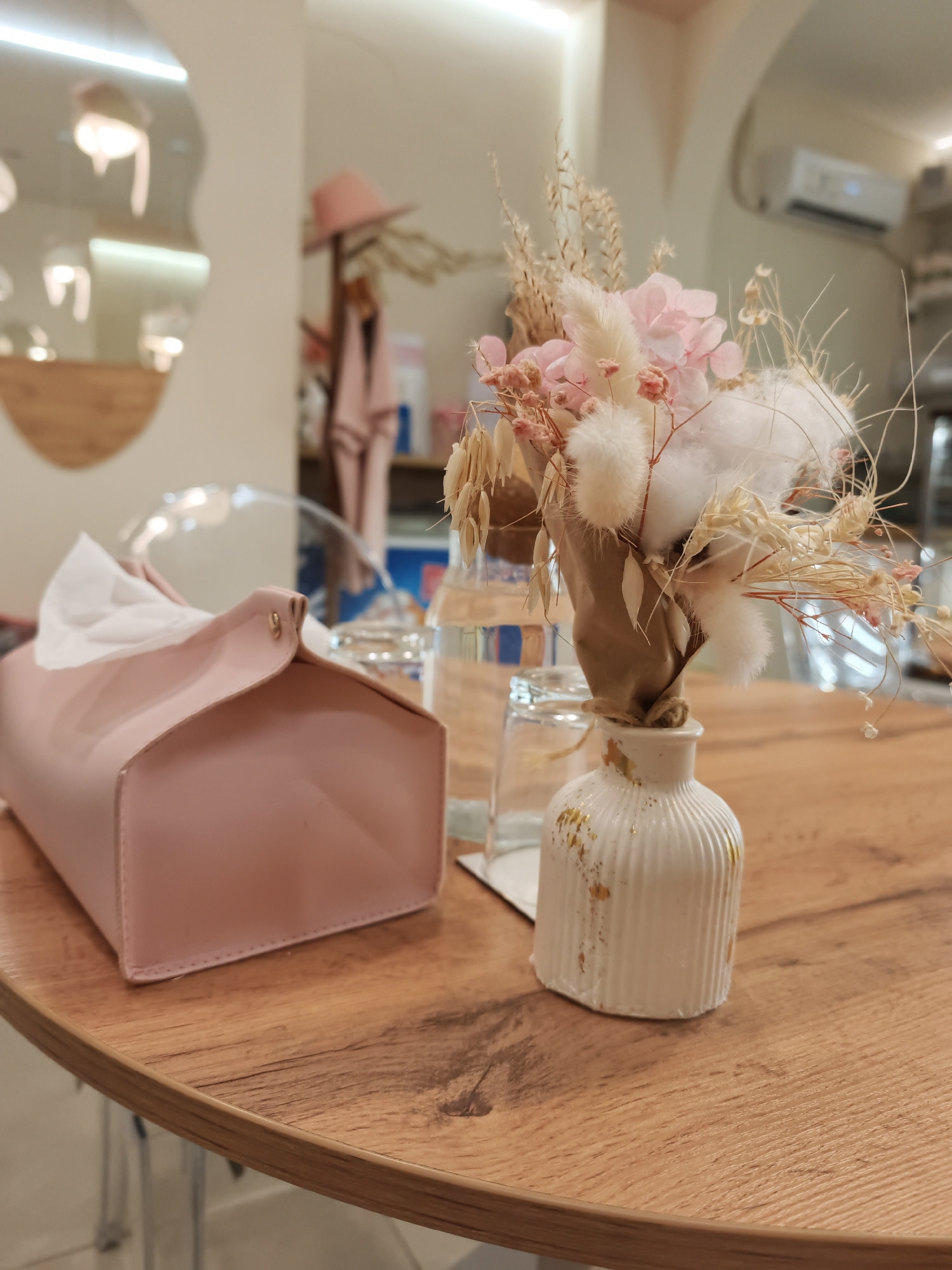 A close-up shot captures a wooden table surface in a dimly lit room. On the table, a pink tissue box with white tissues peeking out is prominently featured on the left. To the right of the tissue box sits a small, white ribbed vase adorned with gold flecks, holding a dried floral arrangement of pink flowers, cotton-like puffs, and wheat stalks. Behind these items, a clear glass of water is visible, along with the blurry reflection of lights and decor on a wall. The overall scene suggests a cozy indoor setting.