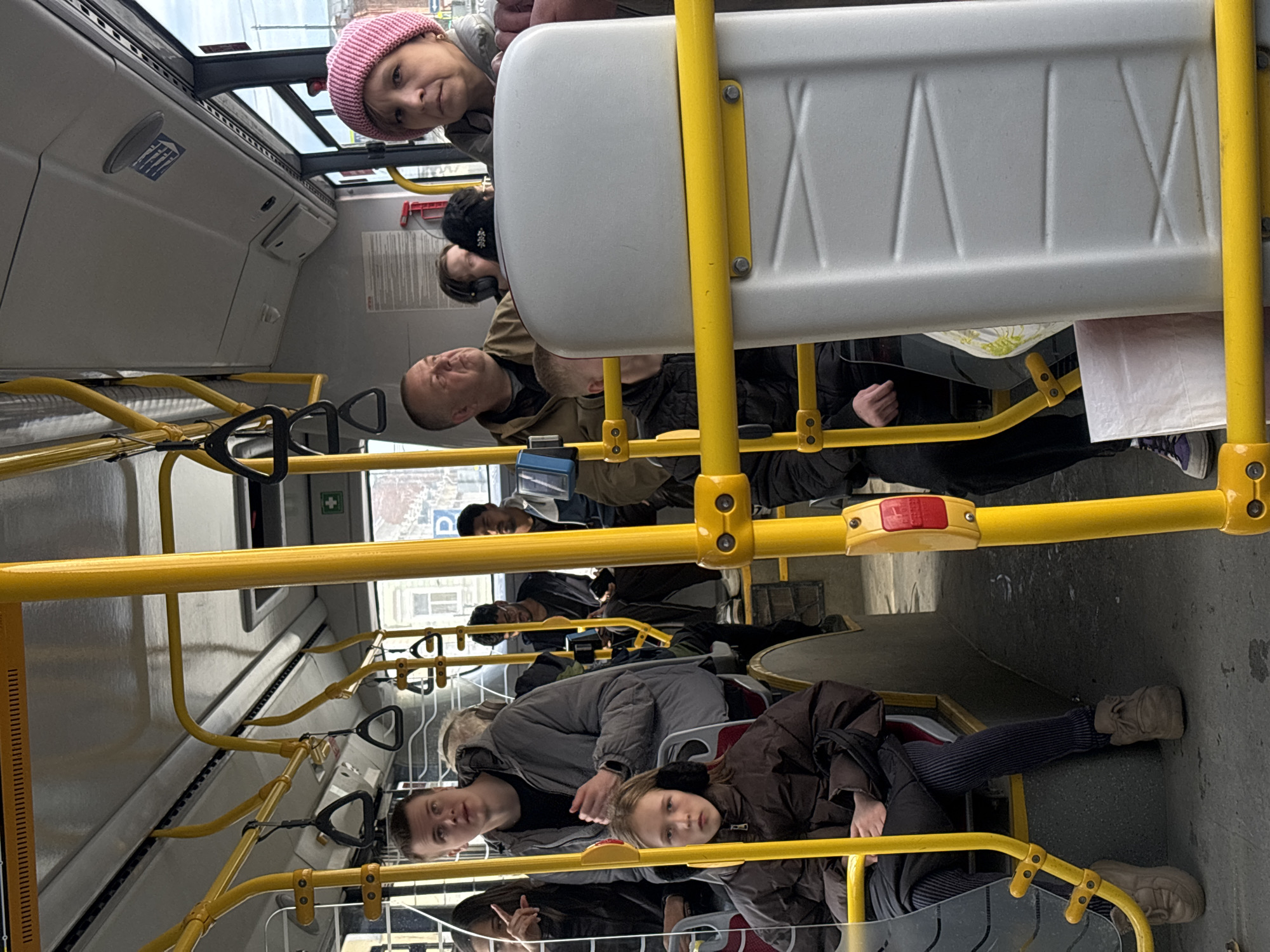 The image shows the interior of a public bus in Perm, Russia. Passengers, including women, men, and children, are seated or standing. The bus features yellow handrails and gray seating. Passengers wear various outerwear, including jackets and hats. A woman in a pink hat is near the front of the bus. Most passengers appear to be looking down or ahead. Buildings are visible through the bus windows.