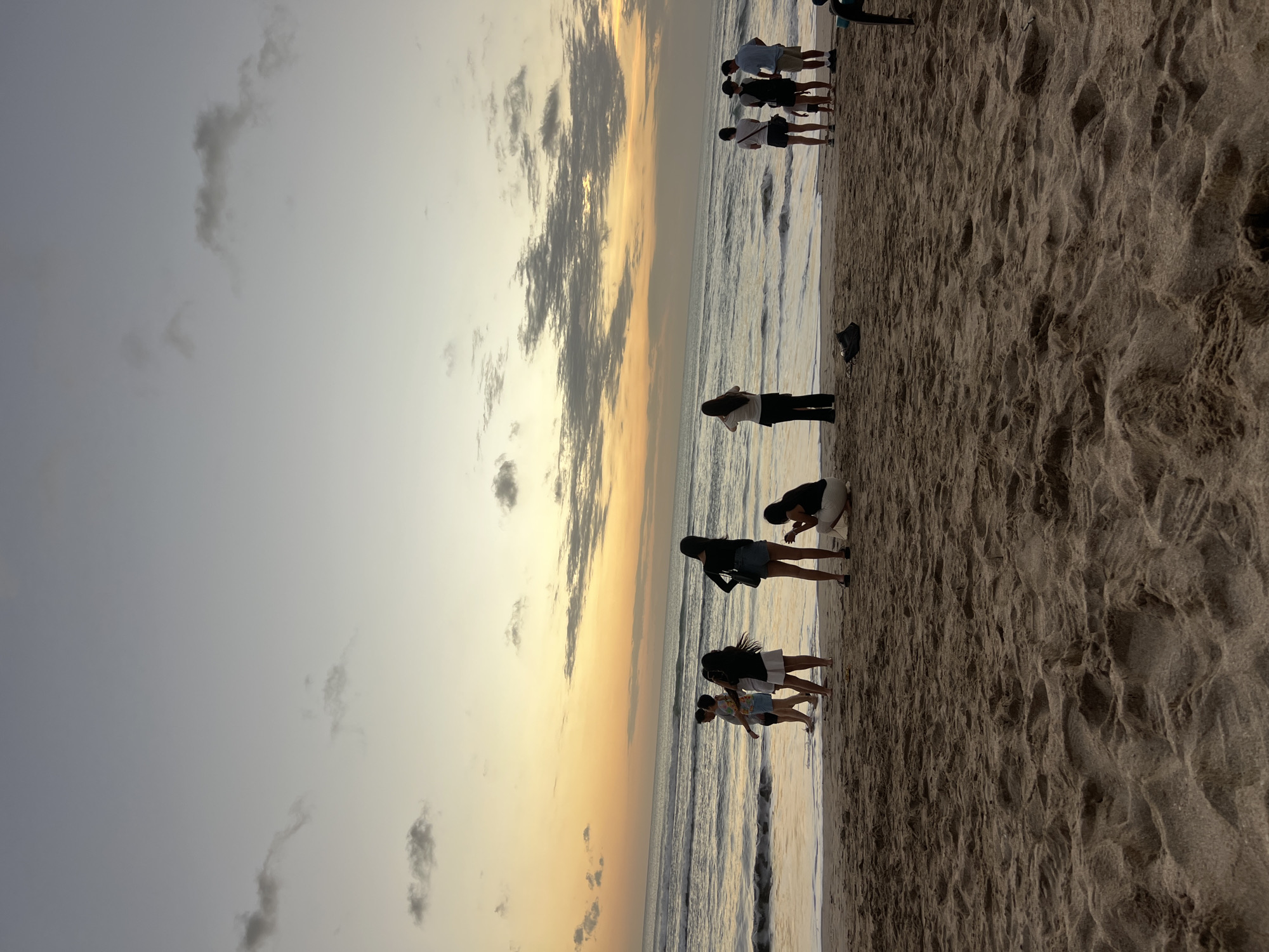 A group of people are standing on a sandy beach in Kuta, Indonesia, near the ocean. The sun is setting, casting an orange glow across the sky and reflecting on the water. Some individuals are standing in shallow water, while others are on the dry sand. One person is crouched down, appearing to take a photo. The sand is textured with footprints and some scattered debris. The overall scene depicts a relaxed evening at the beach.