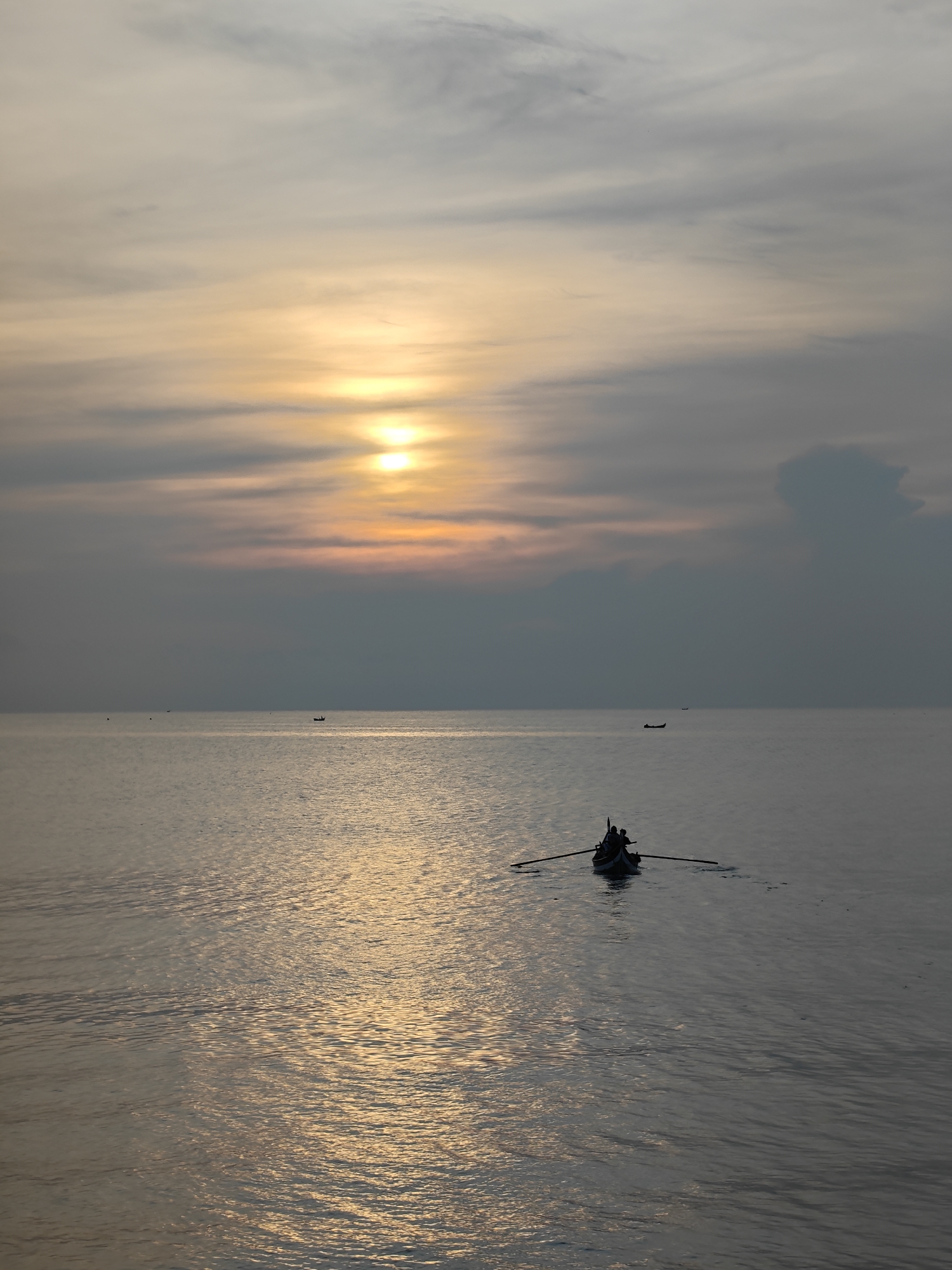 The photograph captures a serene seascape, most likely taken at dusk or dawn, near Lhokseumawe, Indonesia. In the foreground, a small boat, possibly a traditional fishing vessel, is being rowed across the still water. Silhouetted figures can be seen in the boat. The boat reflects the light of the rising or setting sun. The horizon is visible in the distance, punctuated by other small boats. The sky is overcast, with the sun peeking through the clouds, casting a muted glow across the water's surface. The overall mood is tranquil and evokes a sense of daily life at sea.