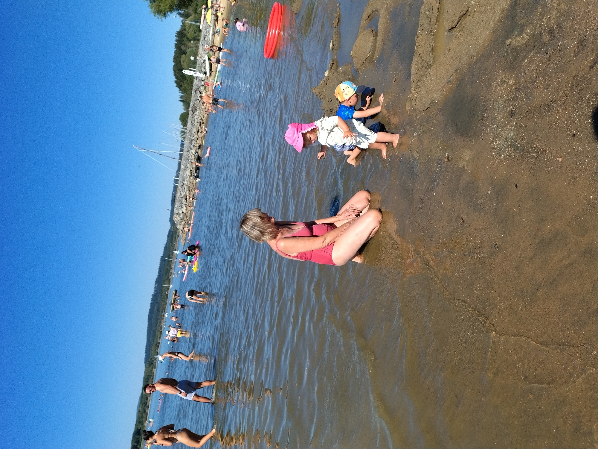 A sunny day at a beach in Radslav, Czechia, shows a woman in a red swimsuit kneeling in shallow water, facing two young children playing in the sand and water. A baby in a white outfit and pink hat stands, while another child in a blue outfit and orange hat crawls near sandcastles. In the background, numerous people are visible in the water and on the shoreline, with boats anchored and sailing further out. The water is calm, and the sky is clear blue.