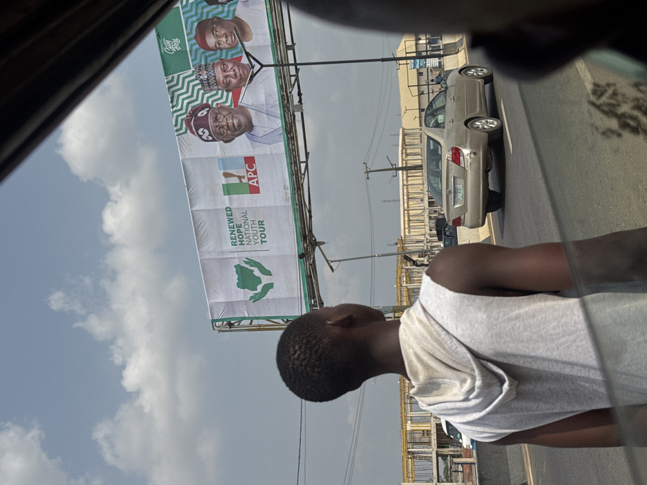The image depicts a daytime scene in Ogbaku, Nigeria, looking forward from inside a vehicle. In the foreground, a young male, seen from the back and right shoulder, stands on what appears to be a road median or shoulder. He has short, dark hair and wears a light-colored sleeveless hooded shirt. His gaze is directed forward.
Dominating the midground is a large billboard displaying political campaign material. The billboard features the text "RENEWED HOPE NATIONAL YOUTH TOUR" and the logo of the All Progressives Congress (APC), including green hands cradling a map of Nigeria. Three male portraits are prominently displayed: Bola Ahmed Tinubu in the center wearing a distinctive cap, flanked by two other male figures, likely political associates.
Below the billboard, a multi-lane road is visible. A gold/tan sedan, possibly a Toyota Camry, with visible license plate "PGE 875Y" and minor rear bumper damage, drives away from the viewer on the left lane. Concrete barriers separate lanes. In the background, a large construction site with multi-story skeletal structures is visible. Utility poles and streetlights line the road. The sky is partly cloudy. The scene is illuminated by natural daylight.