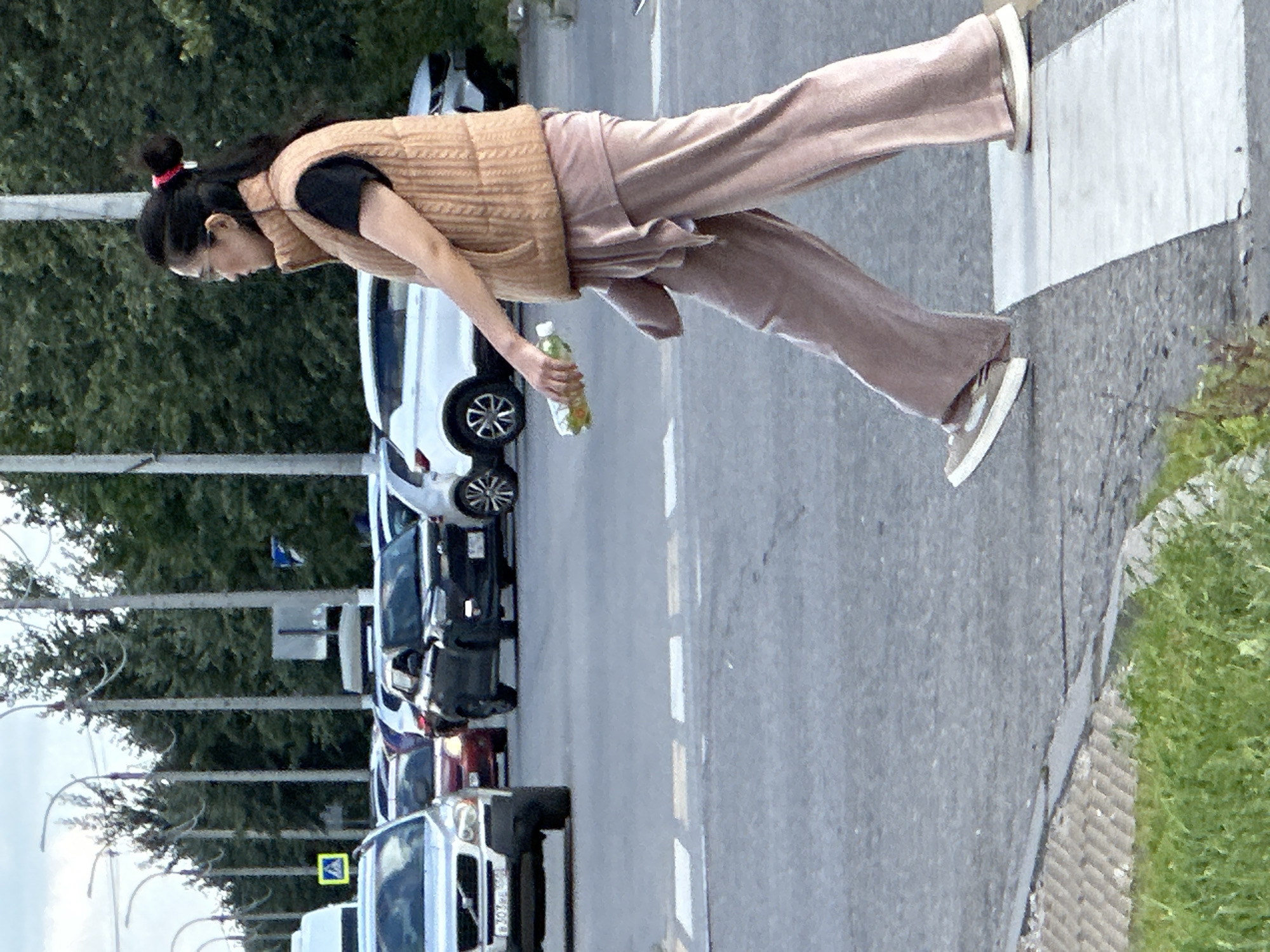 A woman is walking across a pedestrian crossing on a street in Podolsk, Russia. She is wearing a tan puffer vest over a black shirt and loose-fitting pink pants. She is holding a bottle of green liquid in her right hand. Cars are stopped at the pedestrian crossing, indicating that the woman has the right of way. The background shows a line of trees and utility poles. The sky is overcast.