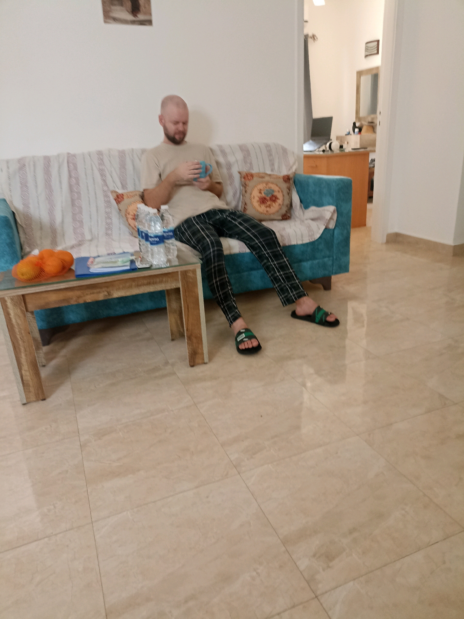 In Al Hadaba, Egypt, a bald man with a beard is seated on a light-colored, striped couch in a living room. He is holding a blue mug in his hands and appears to be looking down at it. He is wearing a light beige t-shirt and dark checkered pants. In front of him, a wooden coffee table holds a bag of oranges, two bottles of water, and a blue binder. The room has light-colored tiled flooring and a white wall. To the right of the couch, a doorway reveals a glimpse of another room with a desk and a laptop. The overall scene is one of a casual indoor setting.