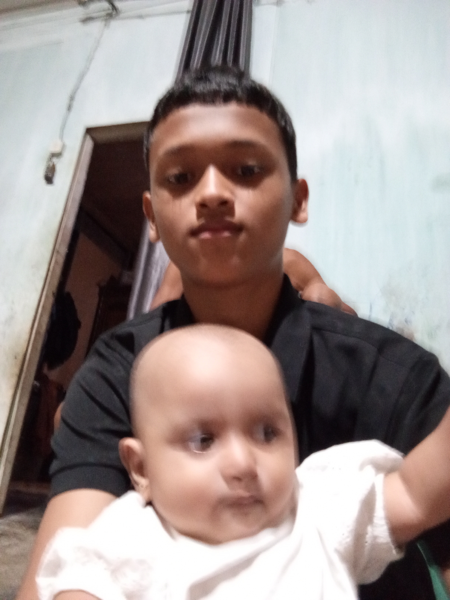 A young boy is holding a baby in a close-up selfie. The boy is wearing a black t-shirt, and the baby is wearing a white top. The background is blurry and appears to be an indoor setting with a wall and a doorway. The lighting is dim.