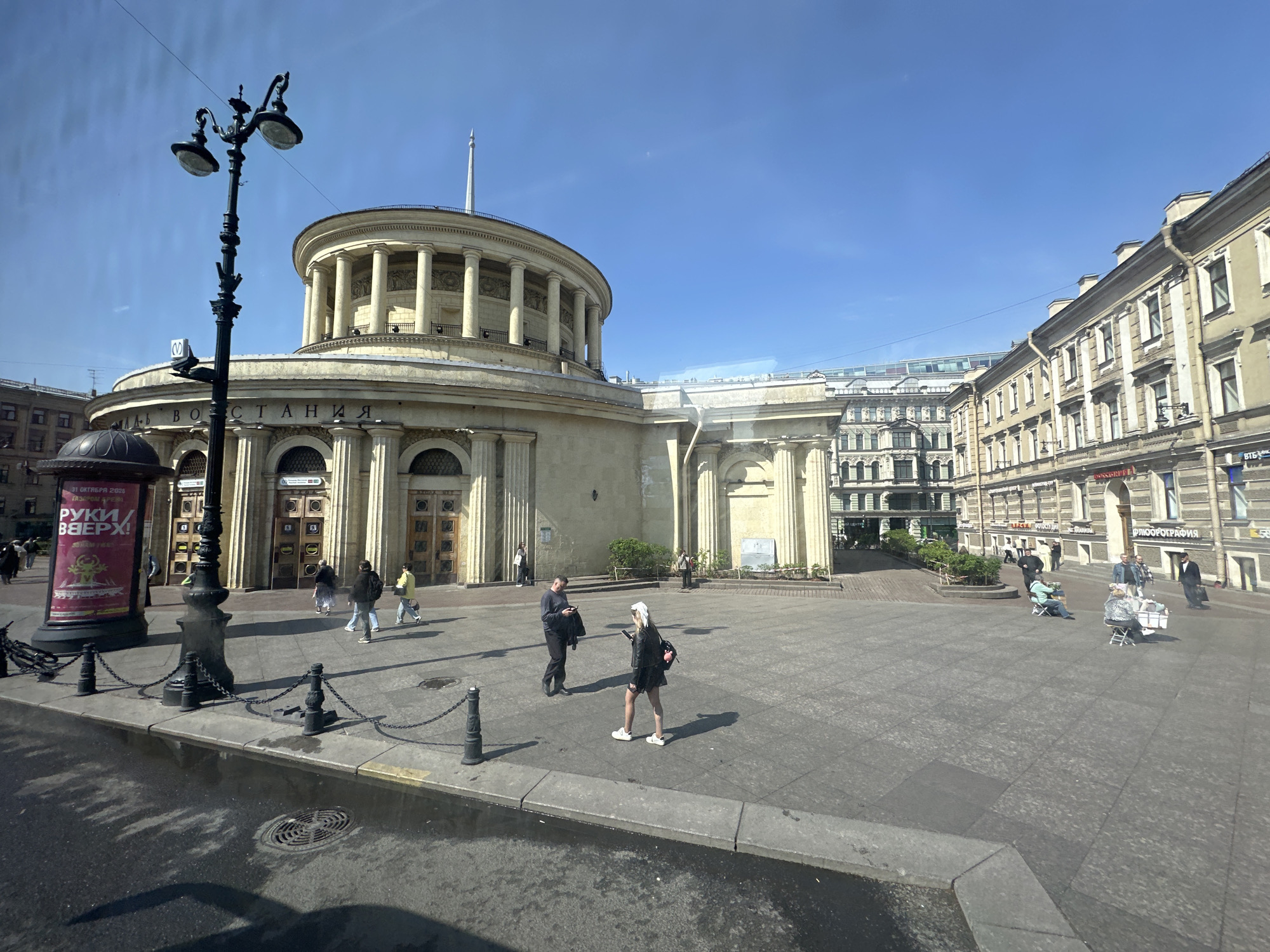 The image depicts a city square in Saint Petersburg, Russia, with the Moskovskaya metro station building prominently featured. The station is a circular structure with a colonnade. People are walking across the paved square, including a woman in a black jacket and white shoes, and a man in a suit and black shoes. A street lamp stands near the edge of the square. A billboard stands near the lamp. There are buildings behind the metro station building. The sky is clear.