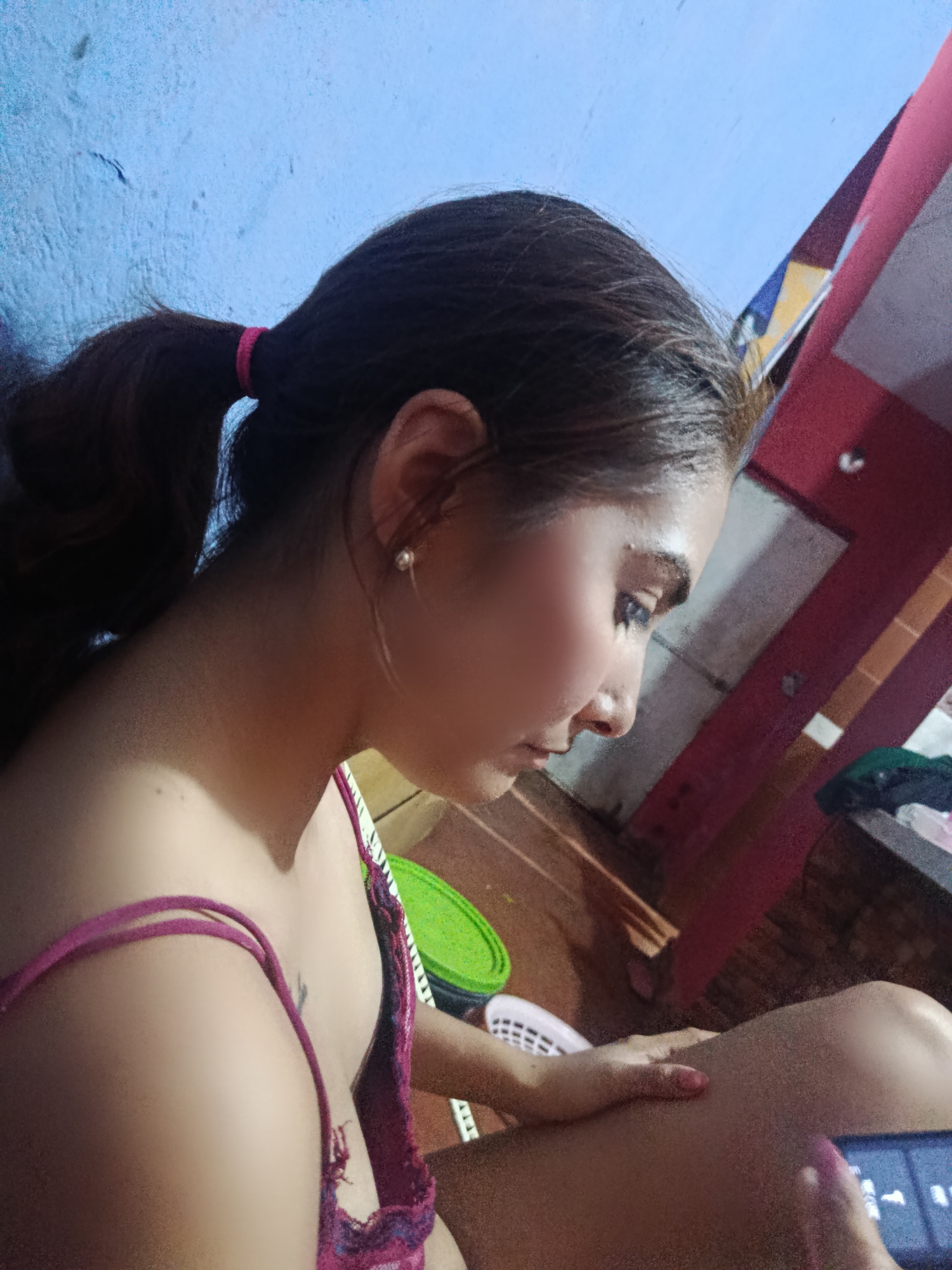 A woman with dark hair pulled back in a ponytail with a pink hair tie is looking down. She is wearing a pink tank top and has a small earring in her left ear. She is sitting with her left hand on her right leg. A phone is visible in her right hand. The background consists of a blue wall, a wooden floor, and a red and white structure. There is no visible information to confirm the location within the city of Juan Griego, Venezuela.