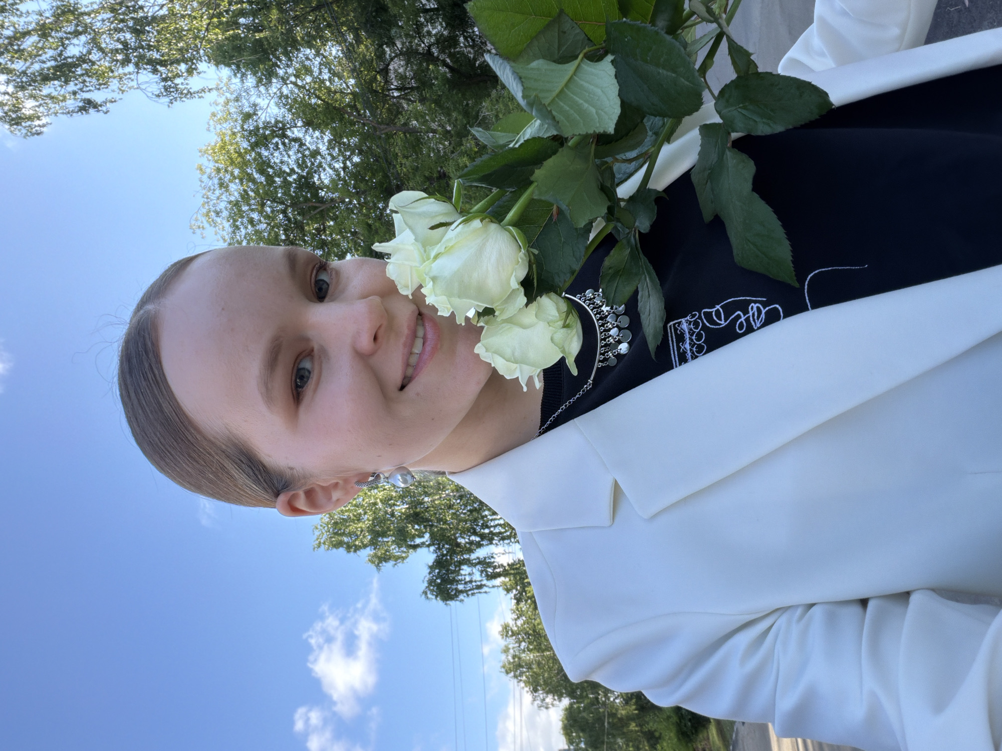A young woman is captured in a close-up, slightly rotated selfie, smiling warmly at the camera. She has light-colored hair pulled back from her face and is wearing a white blazer over a black top. In her left hand, she holds a bouquet of fresh white roses with green leaves, positioned near her face.
Notable accessories include silver-toned drop earrings, each featuring a pearl-like orb, and a silver chain necklace with decorative, reflective elements. The black top features stylized white embroidery, including partially legible text that appears to begin with "De Le" or "De La" followed by an abstract design.
The setting is outdoors under a clear blue sky with a few wispy white clouds. Green tree foliage is visible in the background, suggesting a bright, pleasant day. The woman appears happy and relaxed. The location context for this image is Izhevsk, Russia.
