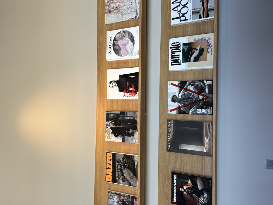 The image displays a wall-mounted magazine rack featuring two parallel, light-colored wooden shelves. The upper wall section is off-white with a warm, circular spotlight illuminating it, while the lower section is painted a light blue-grey. A white electrical outlet with two receptacles is visible on the lower wall, centered beneath the left side of the bottom shelf.
Both shelves are stocked with various international fashion, art, and culture magazines, presented face-out.
On the top shelf, from left to right:
1. A partially visible magazine featuring an individual with white hair, identified as David Bowie.
2. "DAZED" magazine, displaying a man in a plaid shirt reclining on a patterned sofa.
3. "POP" magazine, featuring a woman in dark wide-leg pants and top within an urban setting.
4. "System collections Spring Summer 2020" magazine, showing a woman in a black turtleneck.
5. "AnOther" magazine, with a blonde woman in a pink top lying down.
6. A partially visible magazine featuring a person with light, wavy hair.
7. A largely obscured magazine cover.
On the bottom shelf, from left to right:
1. "Wonderland." magazine, featuring Ellie Rowsell in dark attire seated on a bed.
2. "AKT MAGAZINE", displaying a dark, architectural interior.
3. "V" magazine, titled "MAD LOVE", presenting a black and white image of a man and woman overlaid by a red 'V'.
4. "purple" magazine, showcasing hands and feet with purple floral designs.
5. "LAMPOON" magazine, partially visible, depicting a torso and legs.
6. A largely obscured magazine cover.
7. A largely obscured magazine cover.
No individuals are present in the immediate scene. The location is identified as a minimalist retail environment within the Miami Design District, Miami, United States of America, specifically Dover Street Market Miami, characterized by a curated selection of high-end publications.