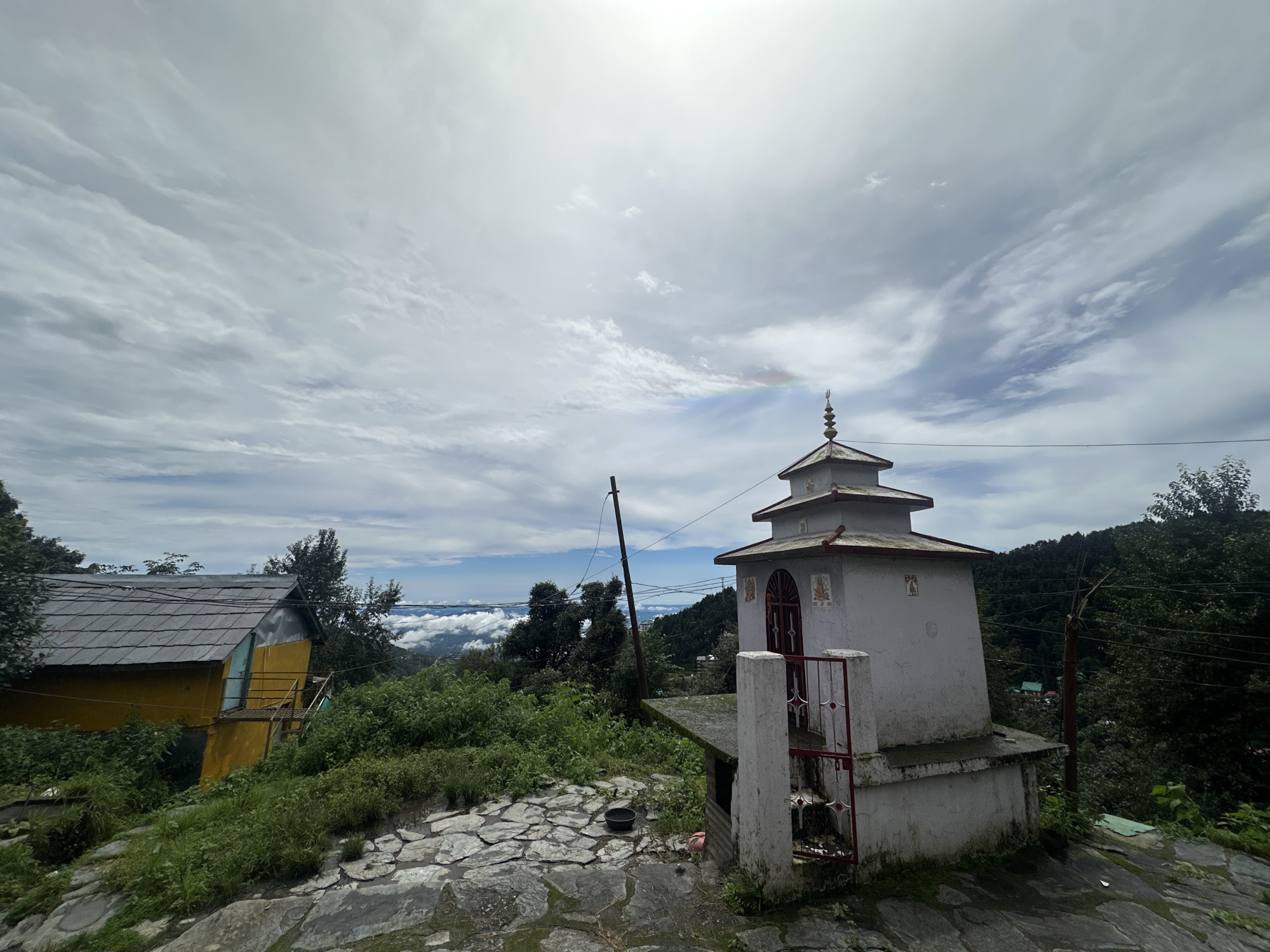 The image shows a view of a small shrine and a house on a hillside, with a cloudy sky overhead. In the foreground, a stone-paved area leads up to a small, white, multi-tiered shrine with a red metal gate. The shrine has a finial on top. To the left of the shrine is a portion of a yellow and blue house with a gray tiled roof and an external staircase. Lush green vegetation covers the hillside around the structures. In the background, the landscape drops away, revealing a valley with clouds and distant trees. The scene is set in Haini, India. There are no people or animals visible in the image. The overall impression is one of a serene, remote location.